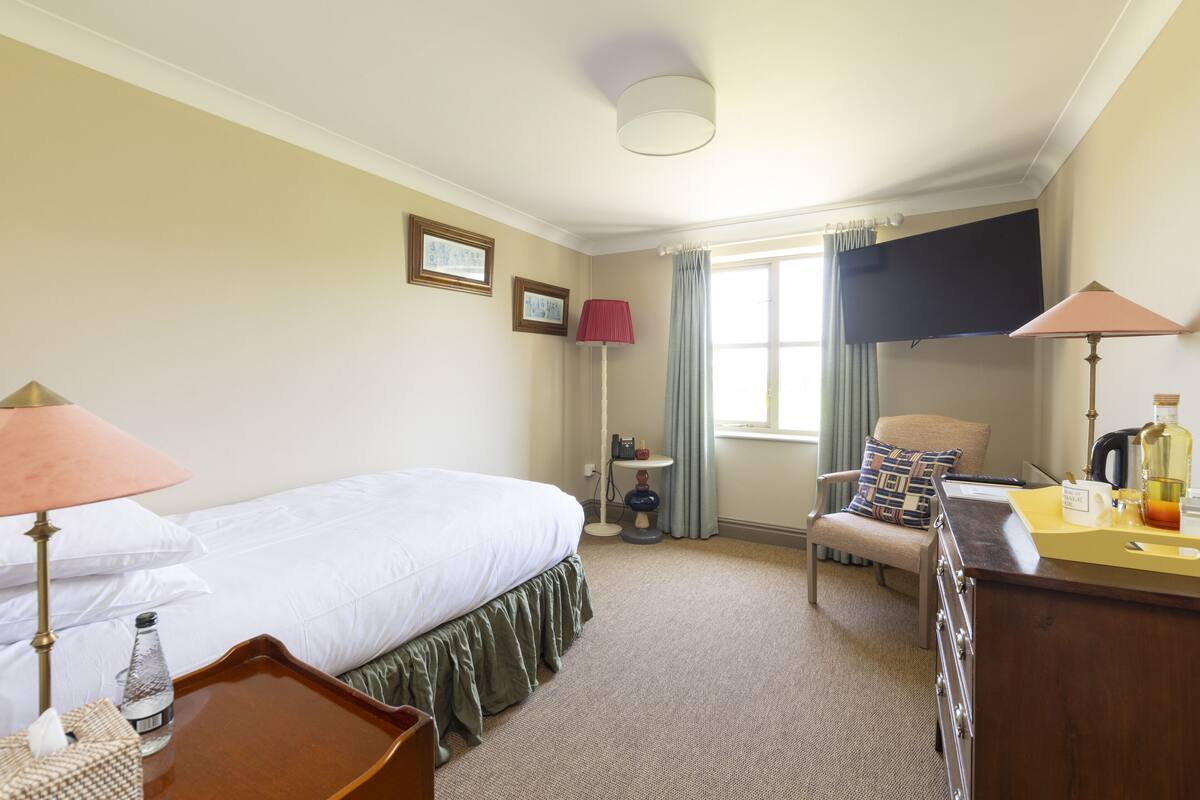 A hotel room with a bed, a desk, a chair, a lamp, a window with curtains, and a small table with a bottle and a cup.
