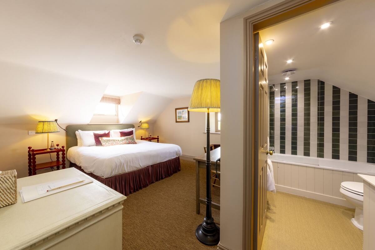 A hotel room with a bed, bedside lamps, a desk, a chair, a lamp, a bathroom with a bathtub, and a toilet.