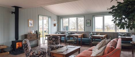 A cozy living room with a fireplace, wooden furniture, and large windows offering a view of the outdoors.