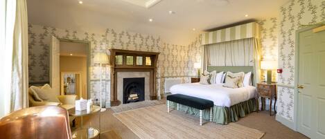 A bedroom with a large bed, a fireplace, a sitting area with a chair and table, and a door to another room.