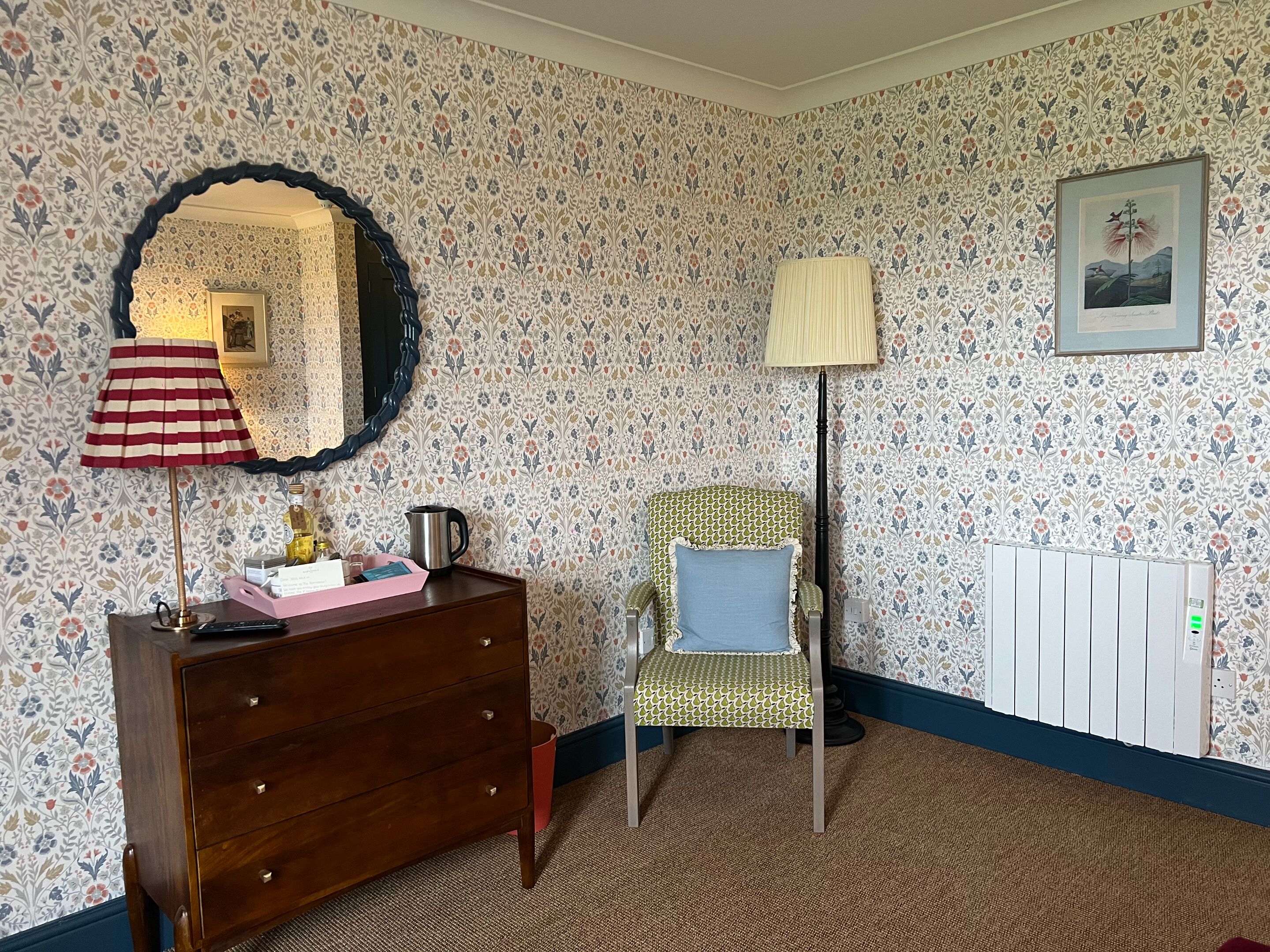 A room with floral wallpaper, a wooden dresser, a patterned armchair, a floor lamp, and a radiator.