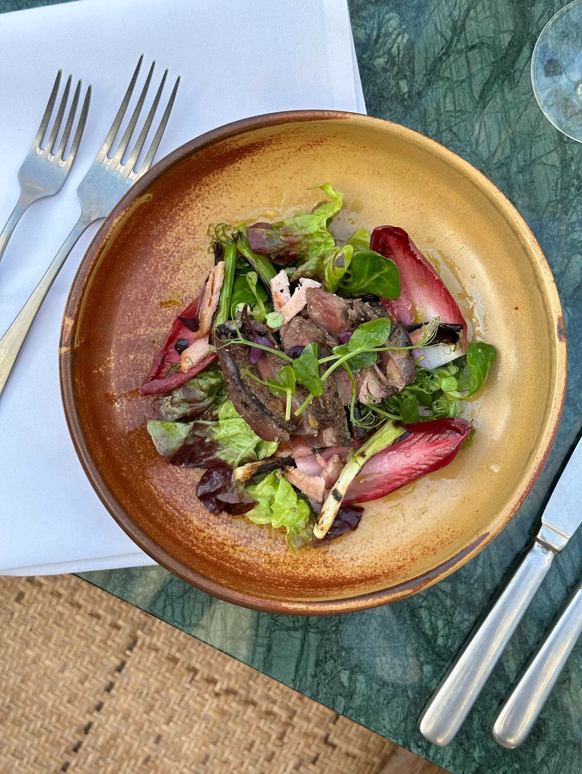 A plate of food with various greens and meat, accompanied by silverware on a table.