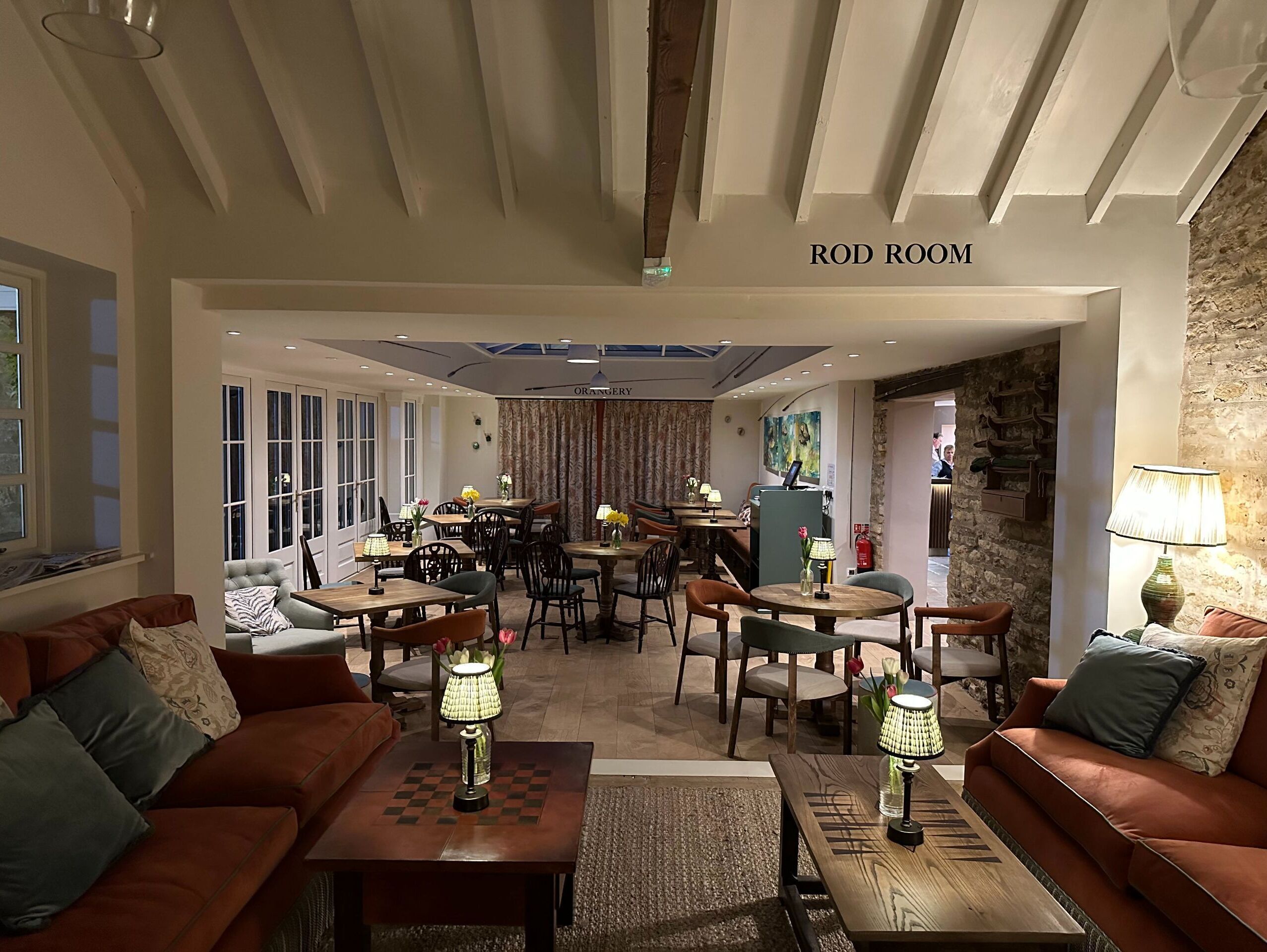 A Rod Room with a cozy seating area featuring a brown sofa and a wooden coffee table, a dining area with wooden tables and chairs, and a stone wall with a fireplace.