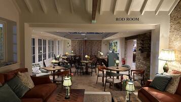 A Rod Room with a cozy seating area featuring a brown sofa and a wooden coffee table, a dining area with wooden tables and chairs, and a stone wall with a fireplace.