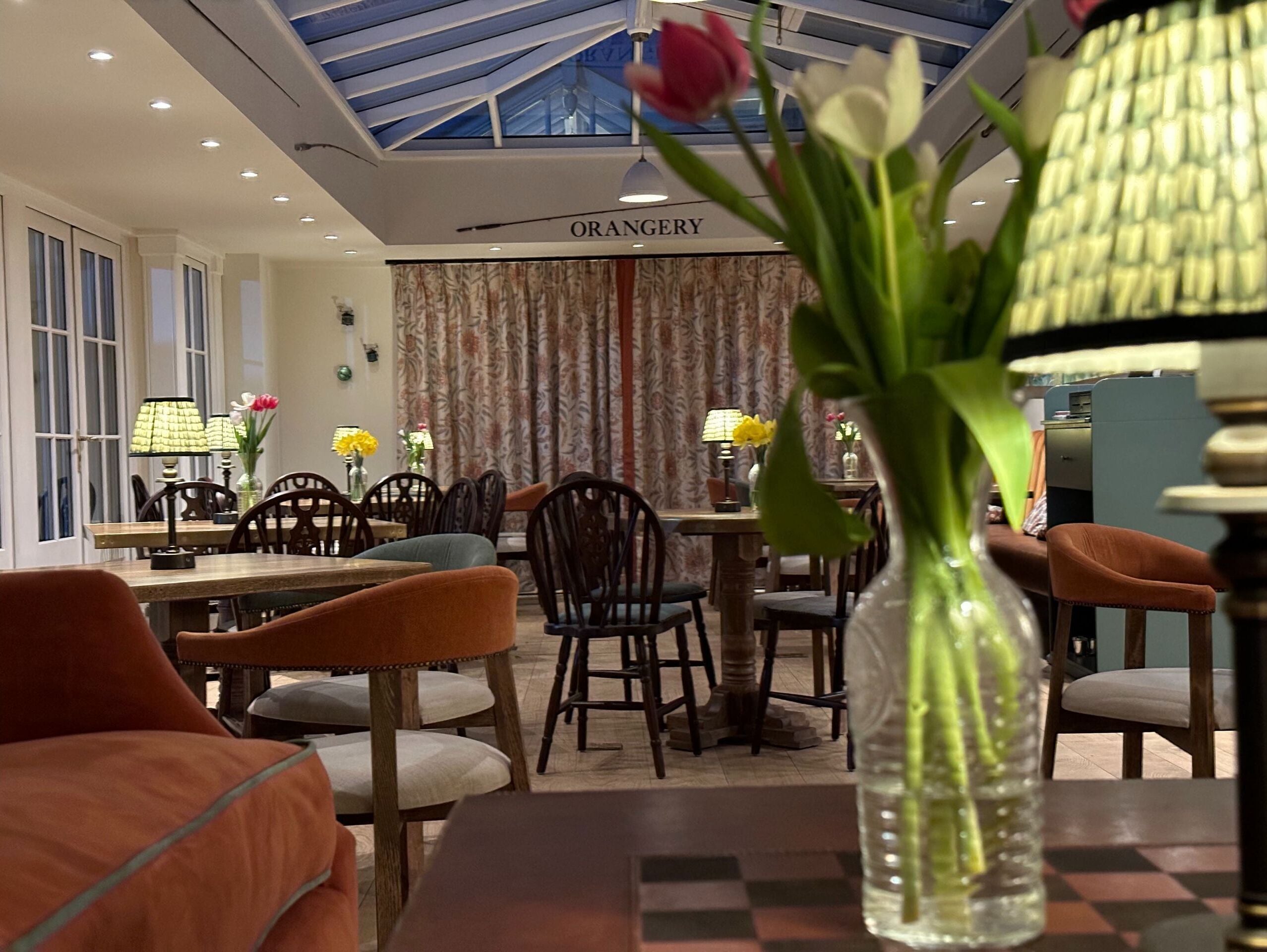 Comedor con mesas y sillas, un jarrón con flores y un letrero que dice 'ORANGERY'.