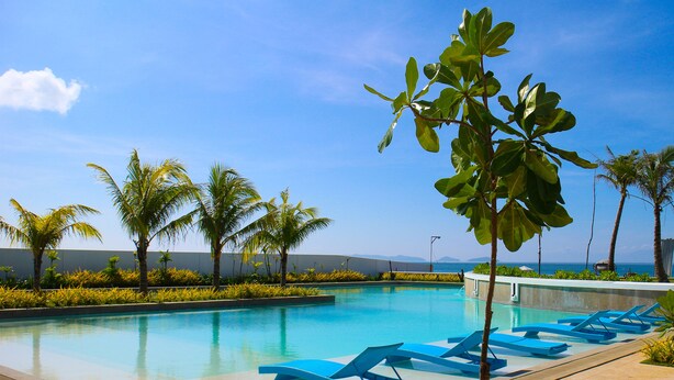 A swimming pool with blue lounge chairs, palm trees, and a clear blue sky. - Ciriaco Hotel and Resort (Calbayog)