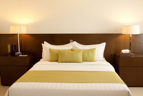 Una cama bien hecha con almohadas blancas y verdes, una manta dorada y dos lámparas de noche sobre mesitas de madera.