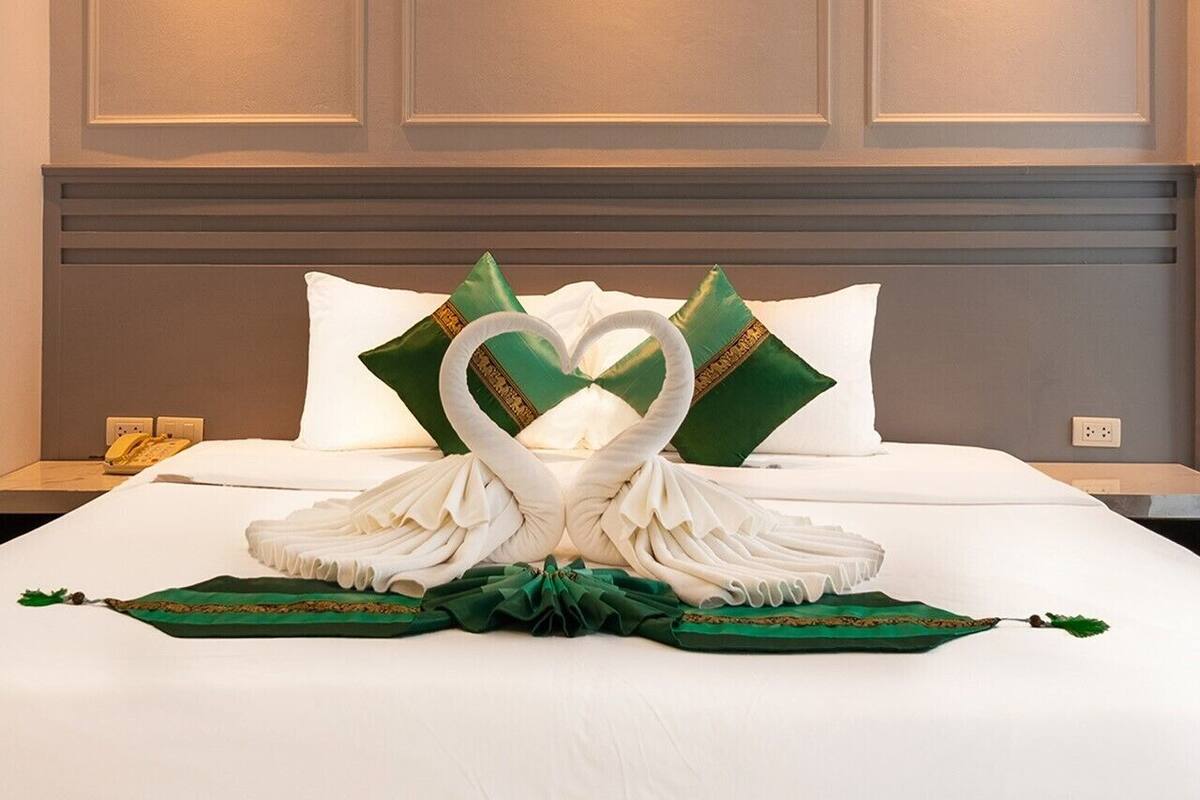 Een net opgemaakt bed met een handdoekarrangement in de vorm van een hart en groene kussens.
