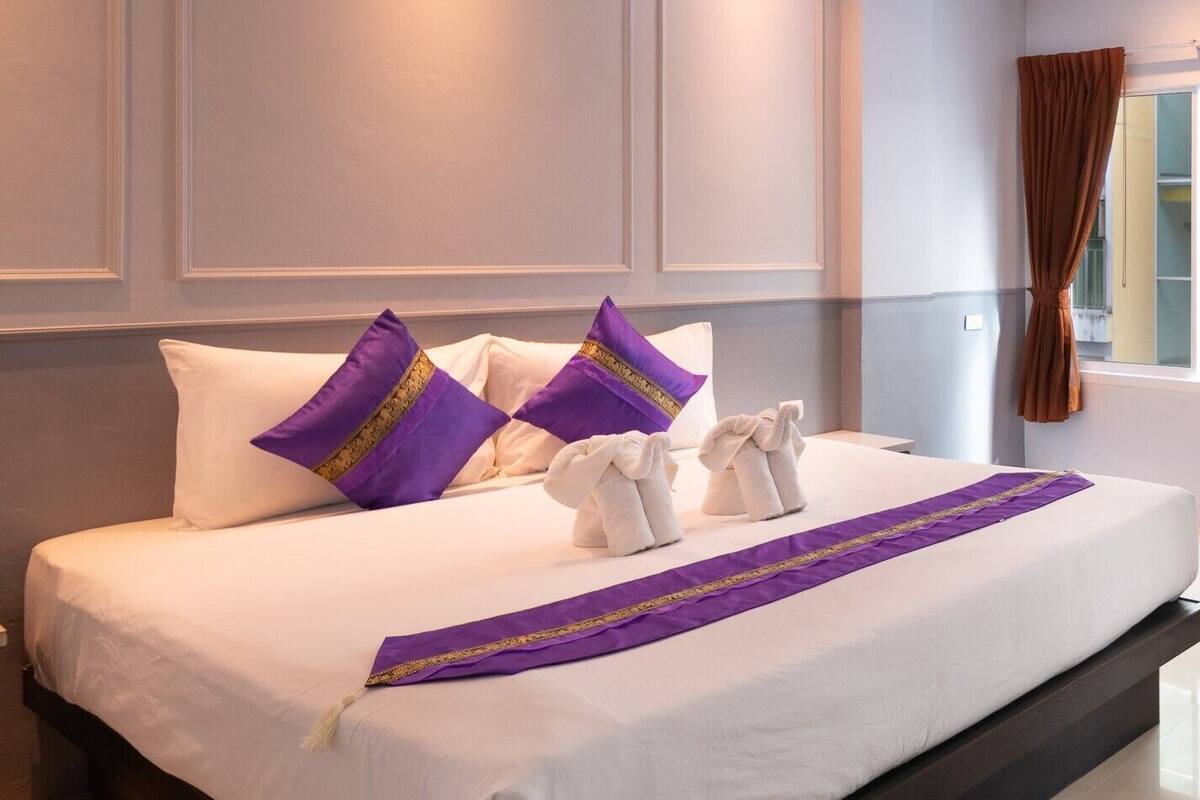Een hotelkamer met een groot bed, witte beddengoed en paarse kussens. Er liggen handdoeken op het bed en een paarse loper.