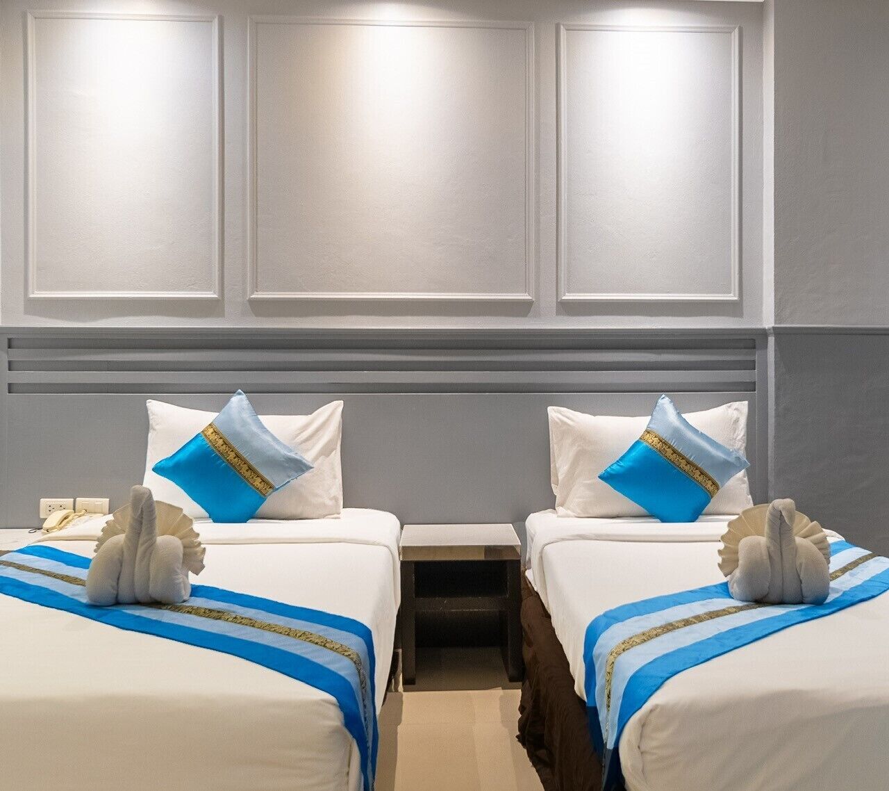 A hotel room with two beds, each with a white bedspread and blue and gold pillows. There is a small table between the beds and a wall-mounted light above.