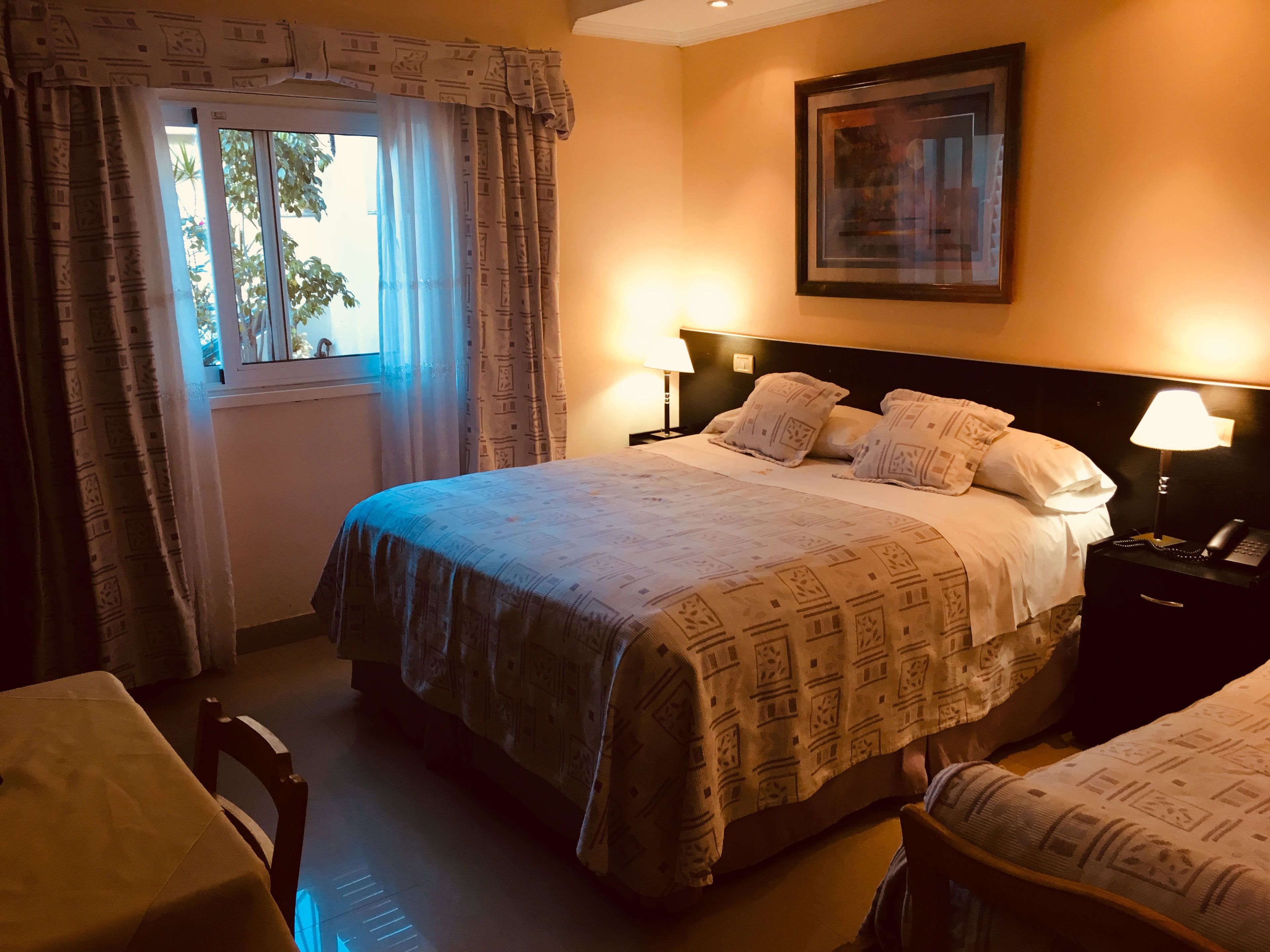 A hotel room with a bed, bedside lamps, a window with curtains, a framed picture on the wall, and a small table.