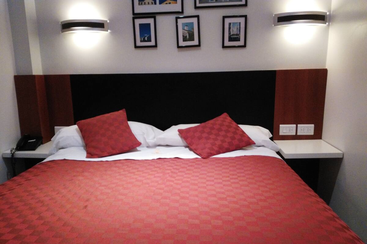 Une chambre d’hôtel avec un grand lit, deux oreillers rouges, une couette blanche et une couverture rouge à carreaux. Il y a deux tables de chevet avec des lampes, un téléphone et un luminaire fixé au mur.