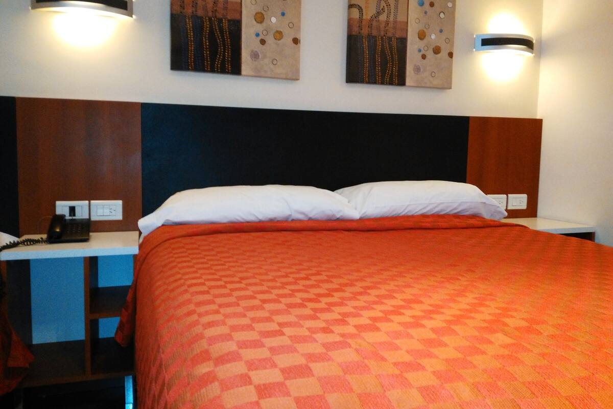 Une chambre d’hôtel avec une couvre-lit à carreaux, une tête de lit en bois, deux appliques murales et un téléphone sur la table de chevet.