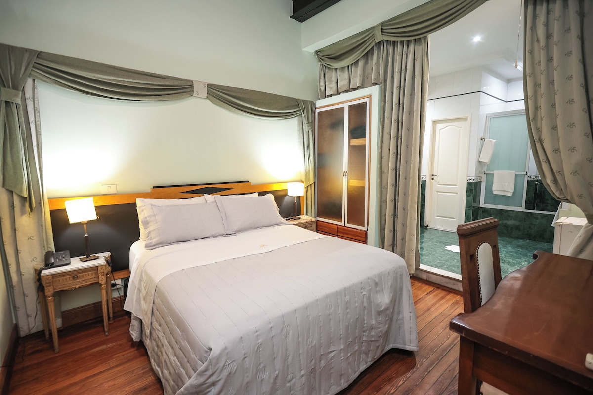 A hotel room with a bed, bedside table, lamp, wooden chair, and a wardrobe.