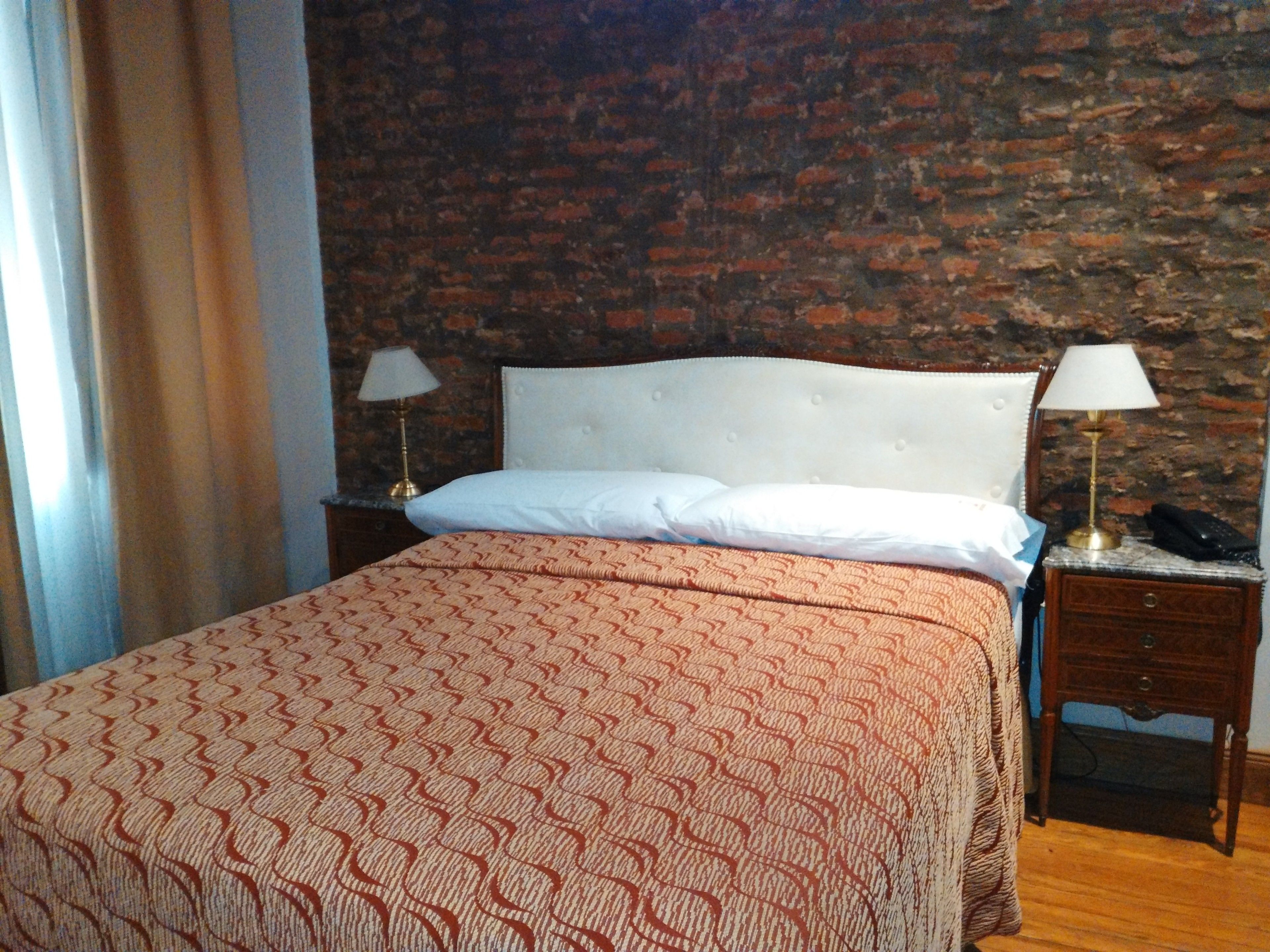A bed with a patterned bedspread, two bedside lamps, a nightstand with a phone, and a brick wall background.