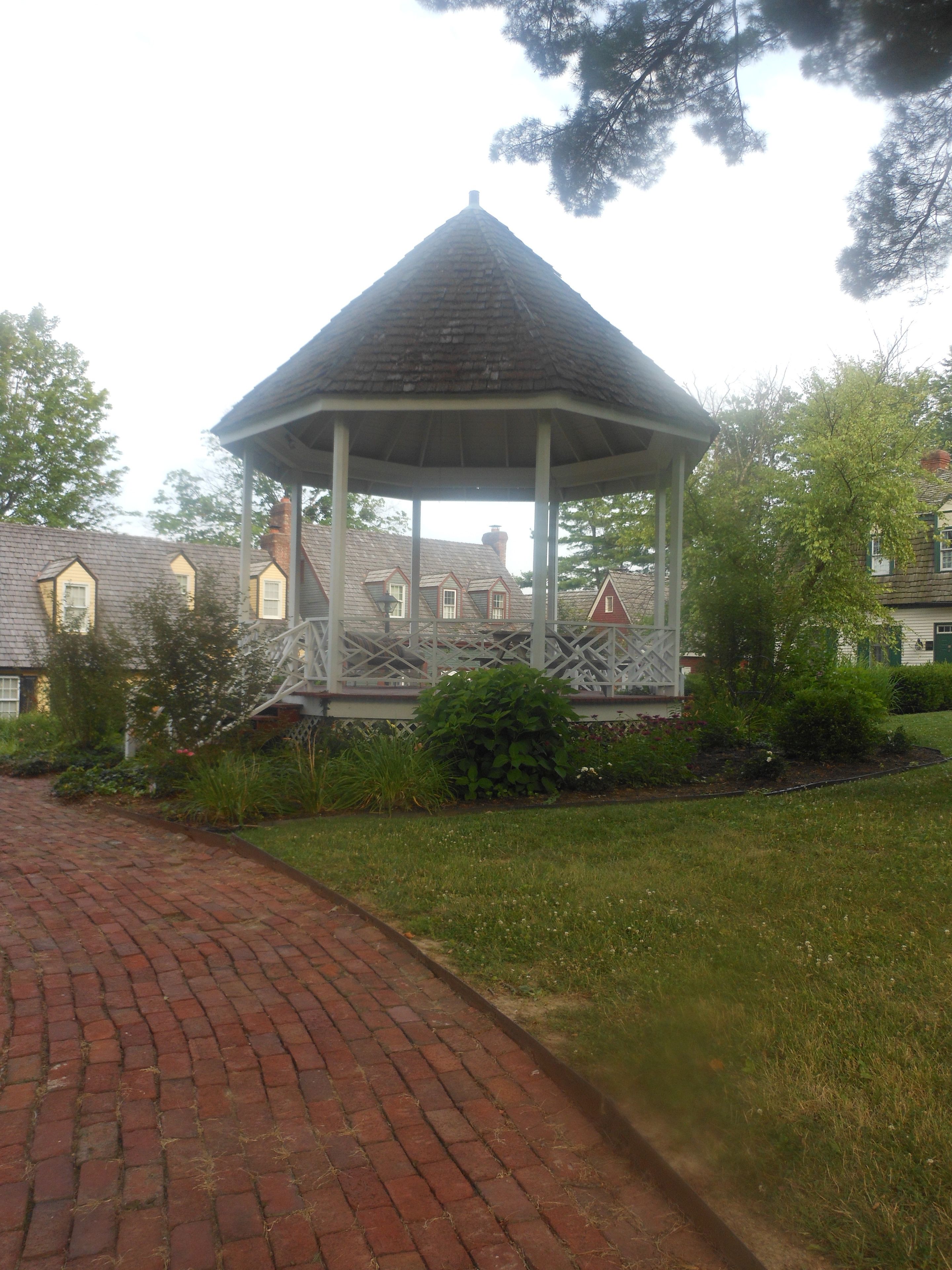 A gazebo with a tiled roof and a white lattice base, surrounded by a brick pathway and greenery.