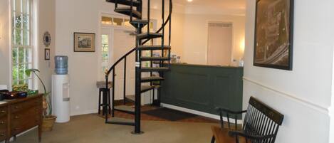 A spacious room with a spiral staircase, a dining area with wooden chairs, a water dispenser, and a green cushioned bench.
