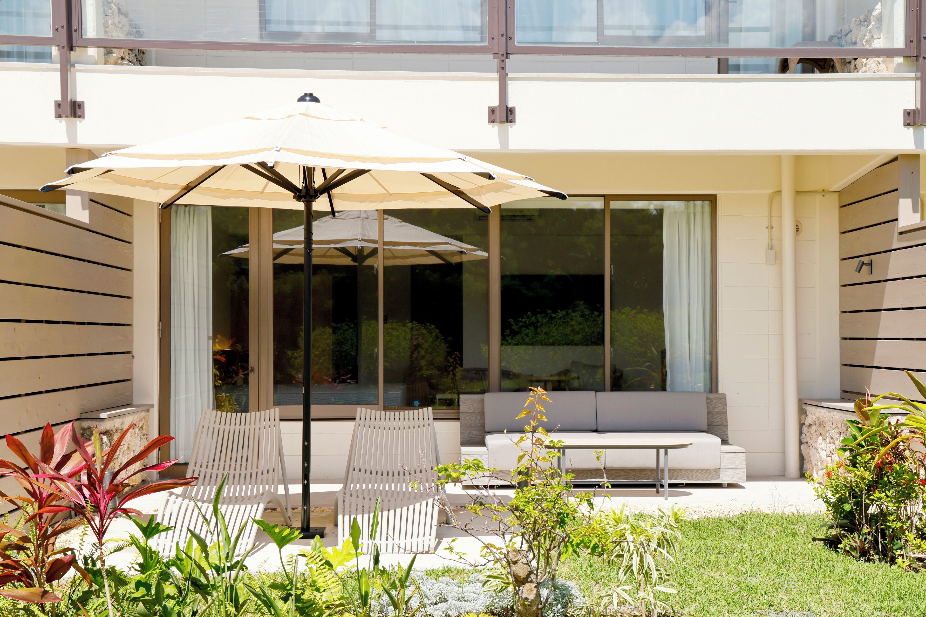 A modern outdoor seating area with a white sofa and two white chairs, an umbrella, and a glass door leading to a pool.