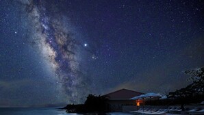 A night scene with a starry sky, a swimming pool, lounge chairs, and a building with a red roof.