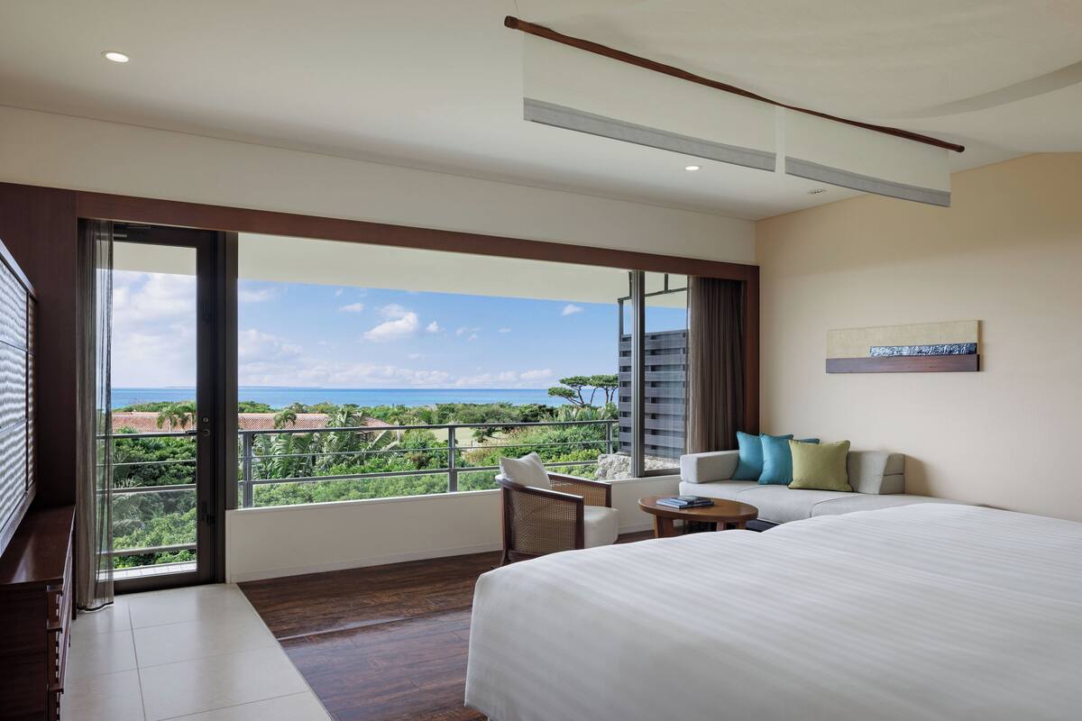 A hotel room with a large bed, a sofa, a chair, and a small table. There is a large window with a view of the ocean and greenery.