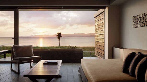 A modern living room with a large sliding glass door leading to a balcony with a view of the ocean and mountains at sunset.