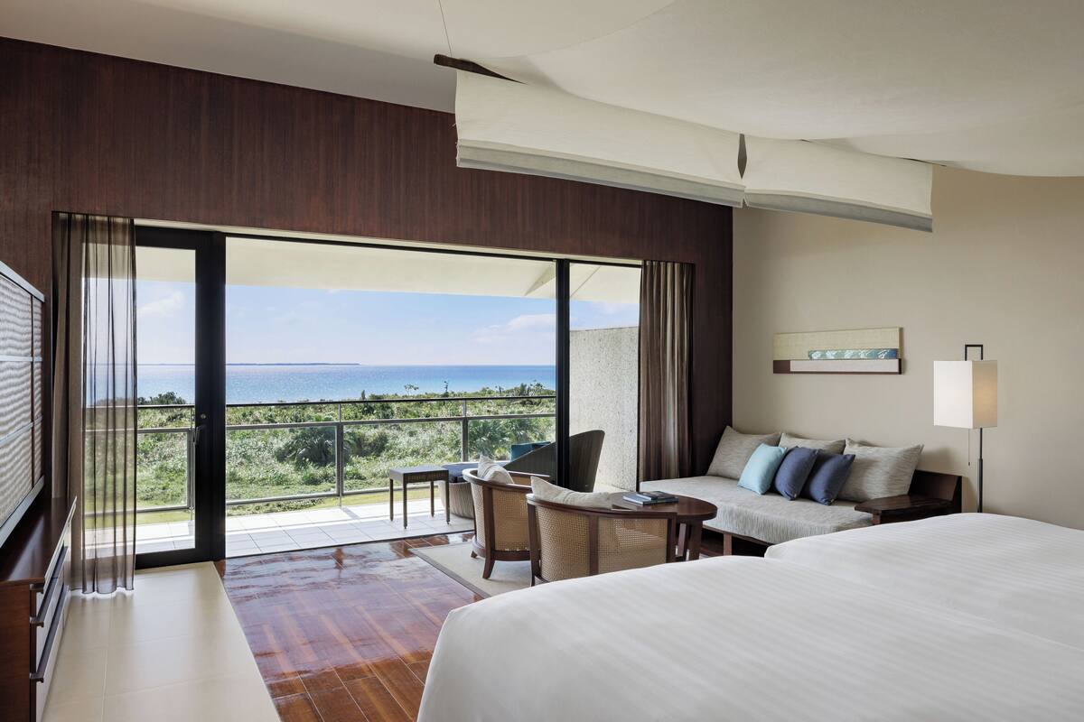 A hotel room with a large bed, a sofa, a dining table, and a balcony with a view of the sea.