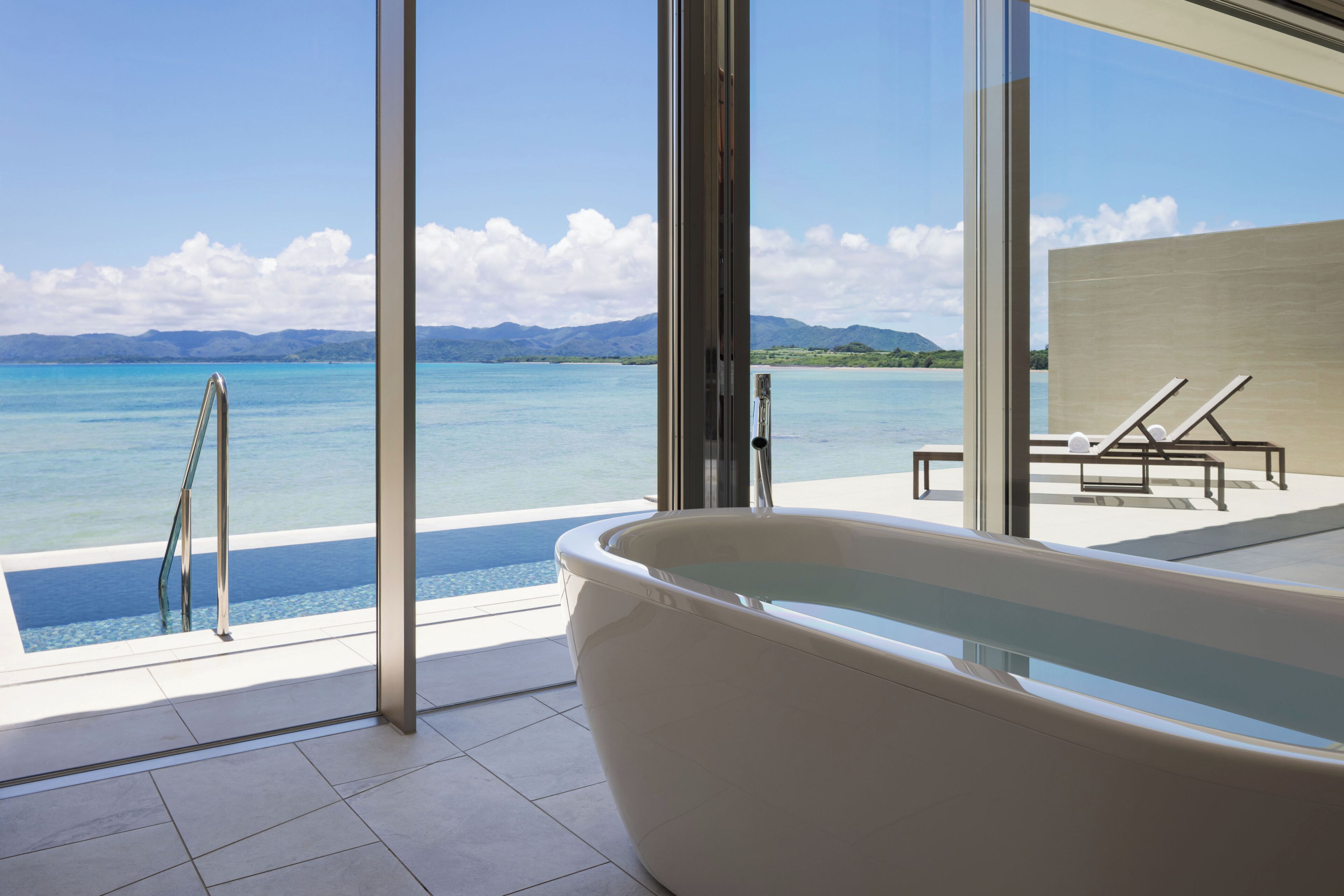 A modern bathroom with a large bathtub, a glass-enclosed shower area, and a view of a clear blue sea and distant mountains.