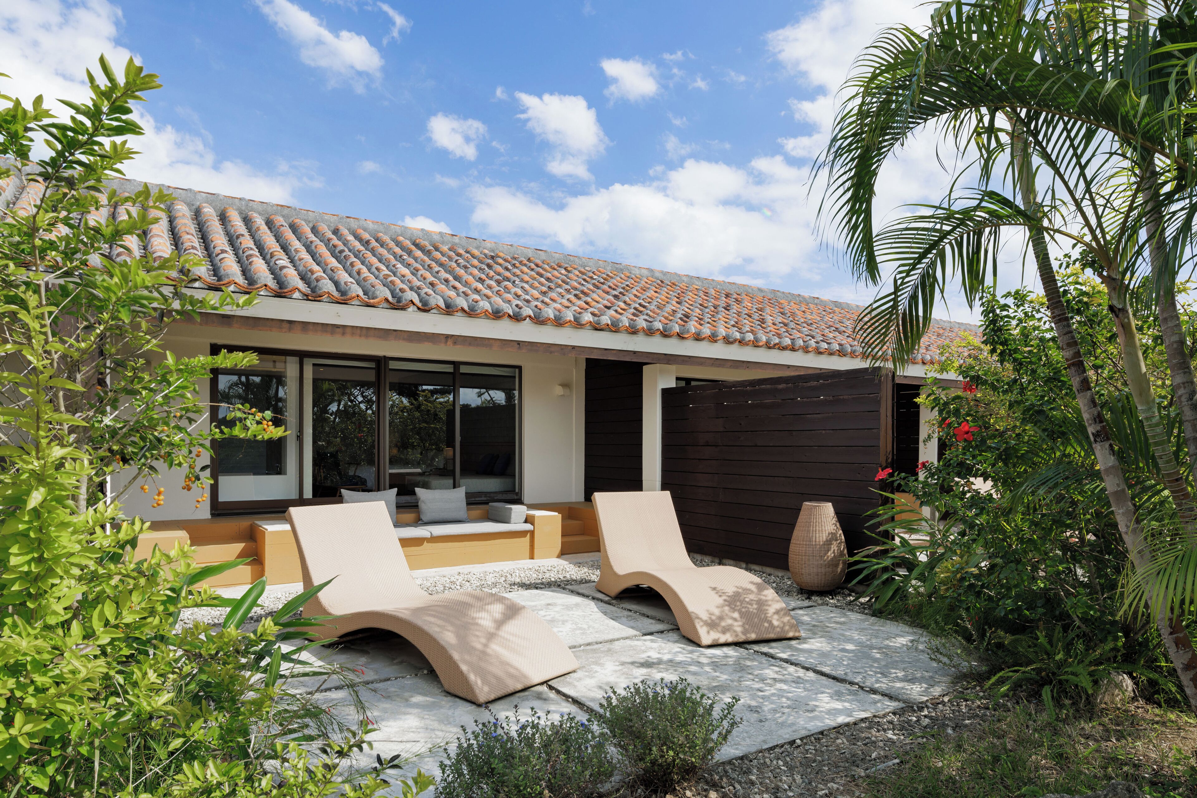 A modern house with a tiled roof, a patio with two lounge chairs, and a garden with various plants.