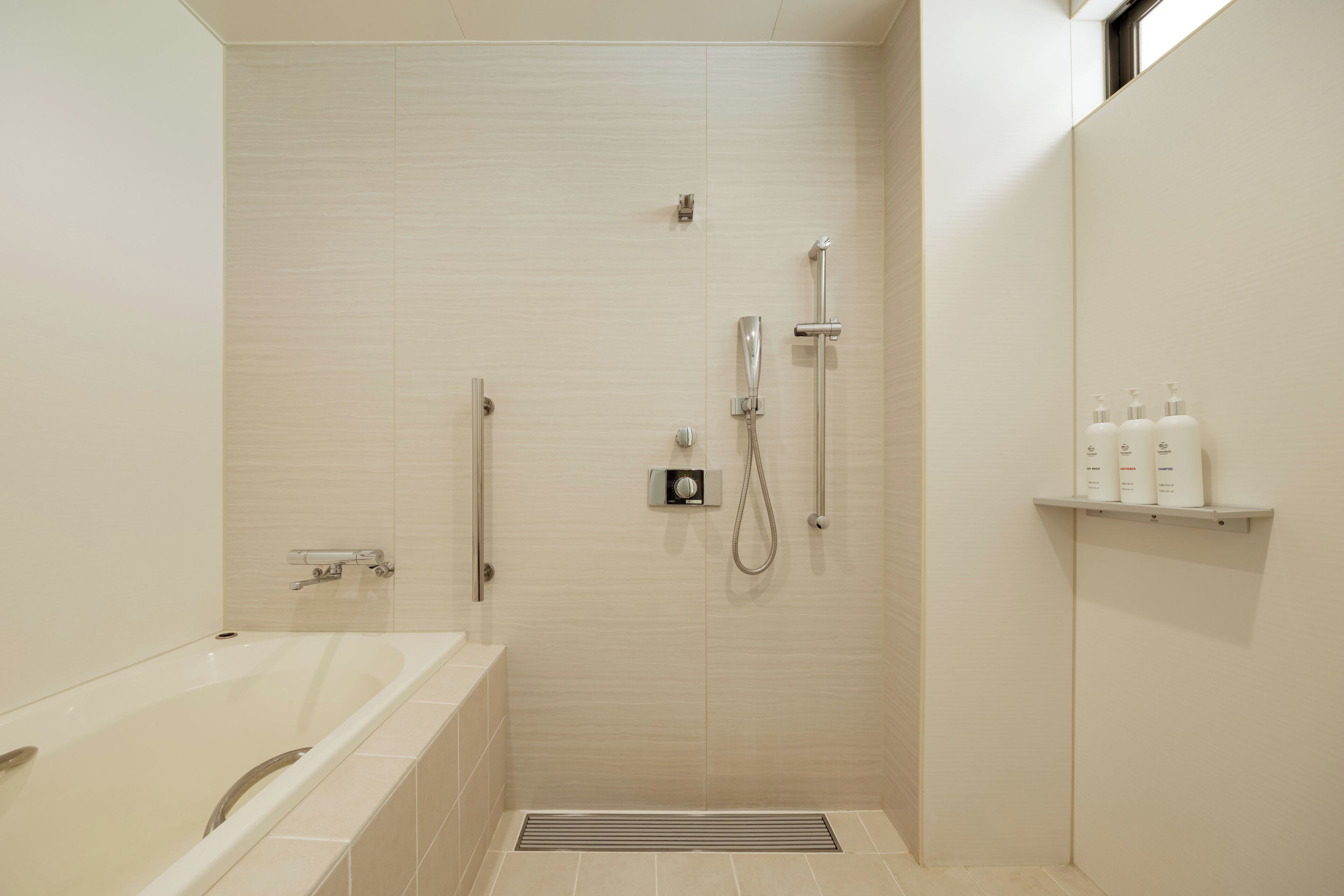 A modern bathroom with a bathtub, a shower with a handheld showerhead, and a wall-mounted faucet. There are three bottles on a shelf above the shower.