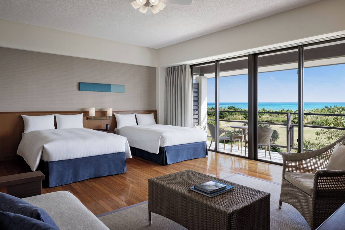 A hotel room with two beds, a balcony with a view of the ocean, and a seating area with a table and chairs.