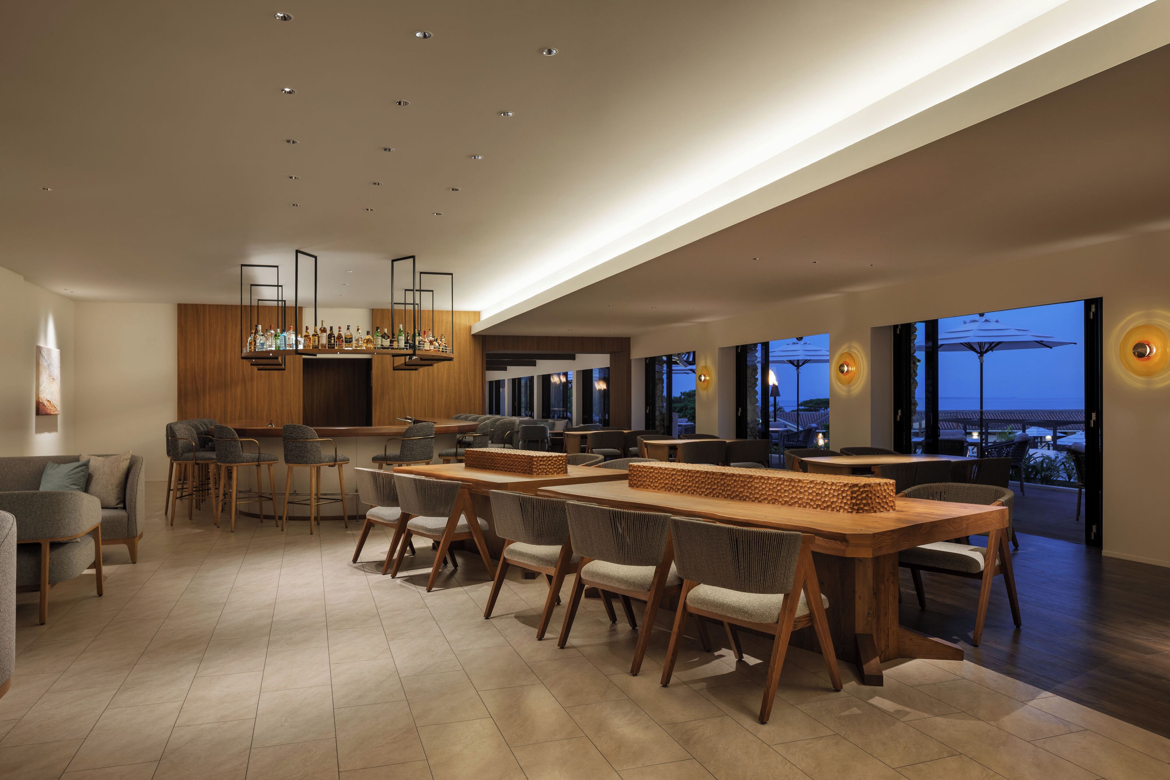 A spacious, modern dining area with high tables and chairs, a bar area with stools, and large windows offering a view of the evening sky.