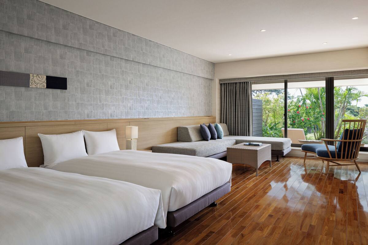 A modern hotel room with two large beds, a sofa, a chair, and a small table.