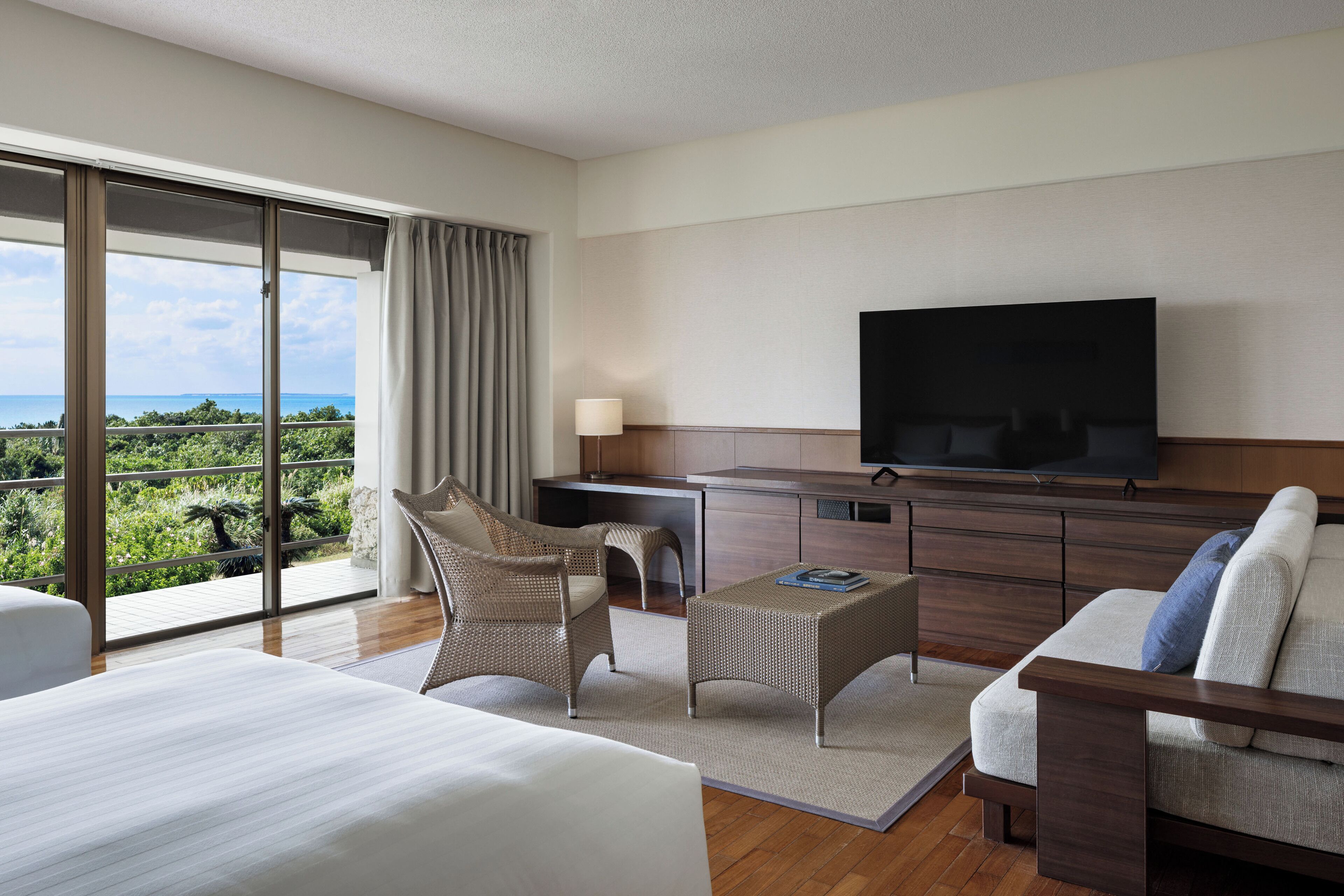 A modern hotel room with a large bed, a sofa, a TV, and a balcony with a view of the sea.