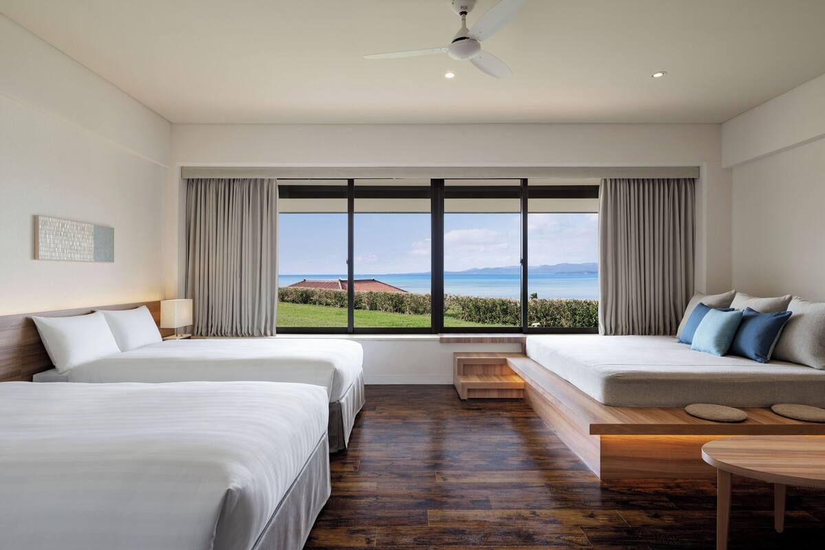 A hotel room with two beds, a sofa, a wooden bench, and a view of the sea.