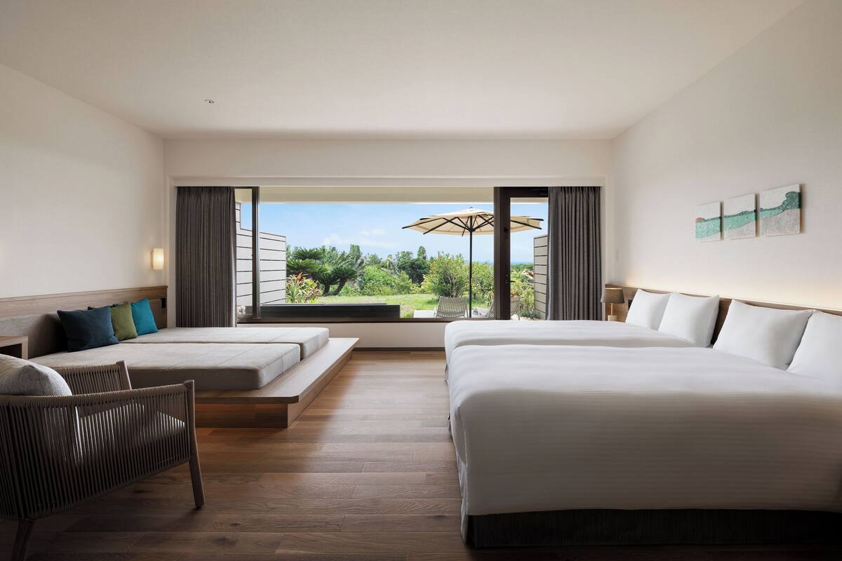 A hotel room with two beds, a sofa, a chair, and a view of a garden.
