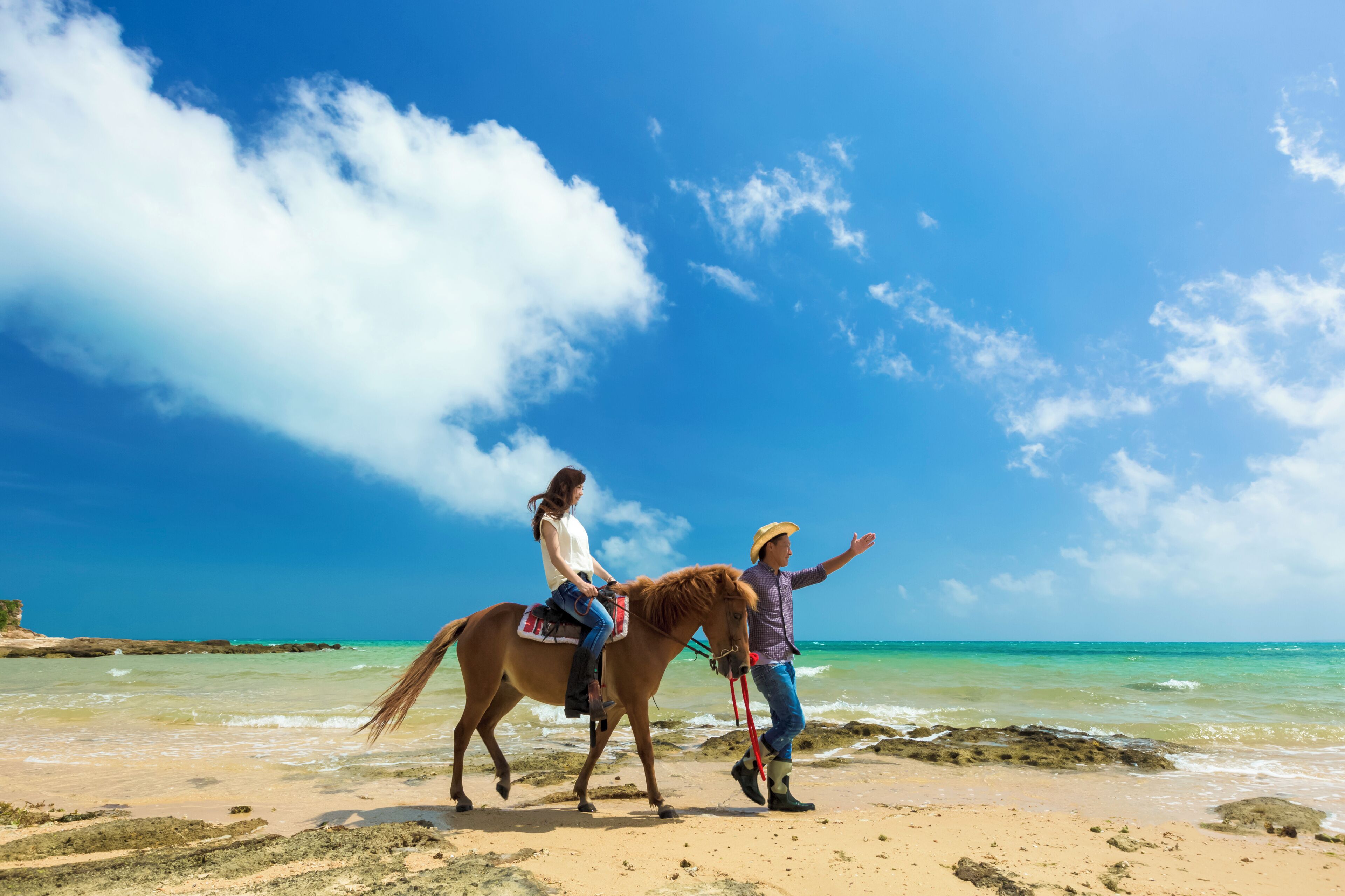 A beach scene with a person leading a horse and another riding it.