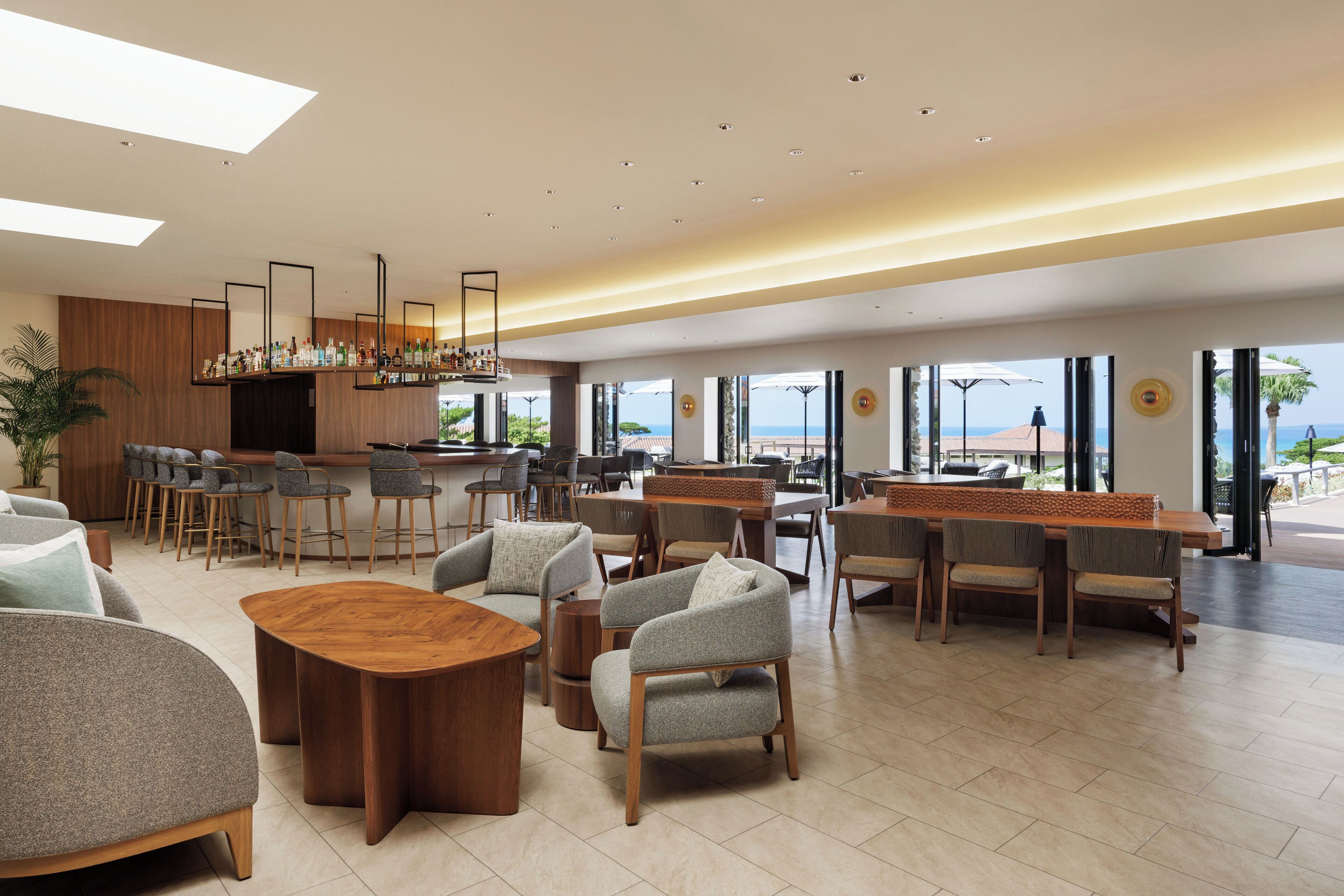Un hall d’hôtel moderne doté d’un bar, de différents espaces de détente et de grandes fenêtres offrant une vue sur la mer.
