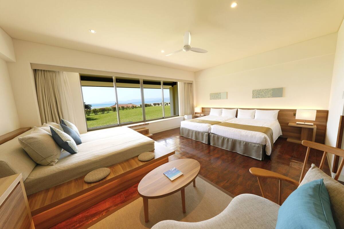 A hotel room with a sofa, two beds, a coffee table, and a view of the outdoors.