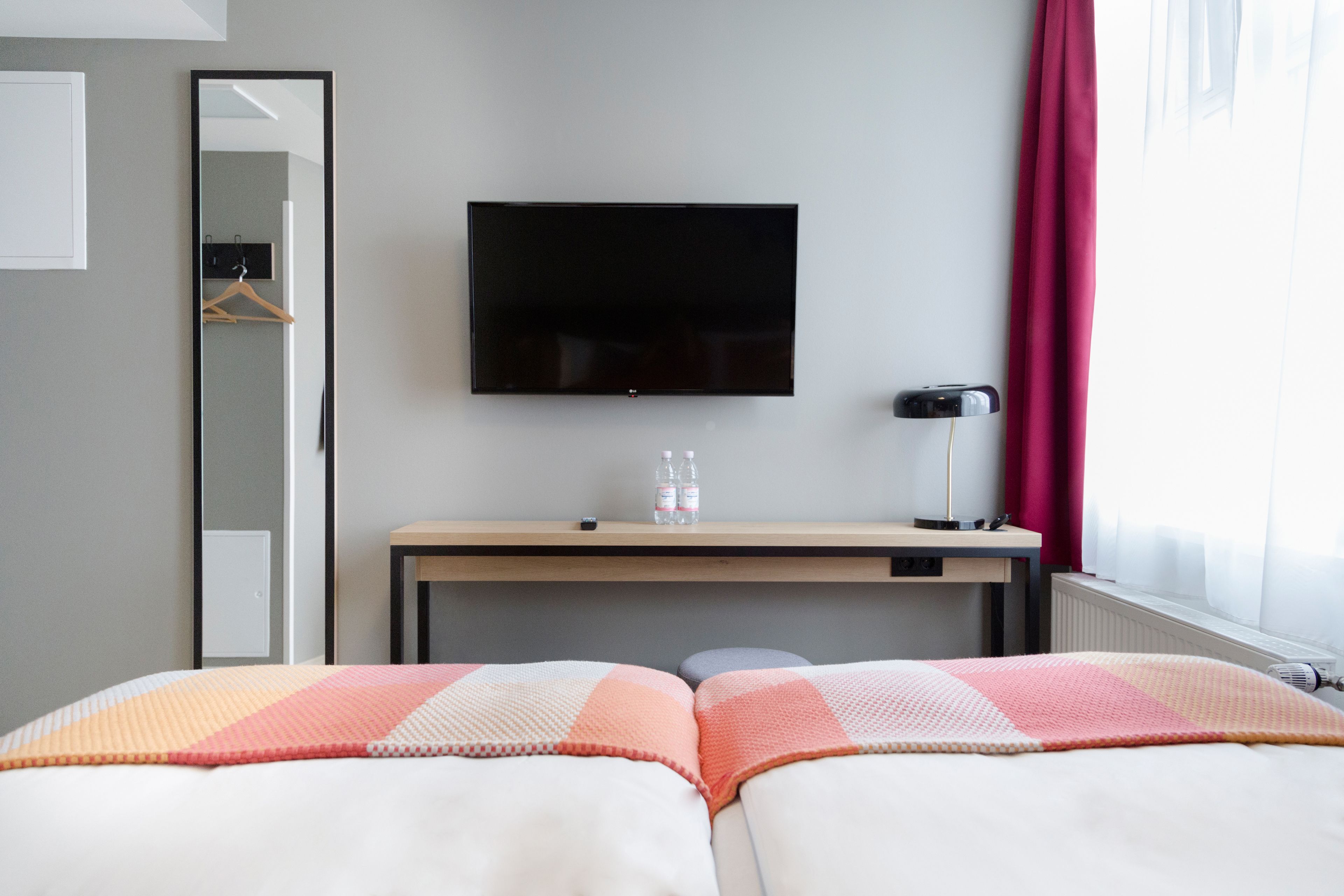A hotel room with a bed, a desk with a flat-screen TV, a mirror, a radiator, and a window with curtains.