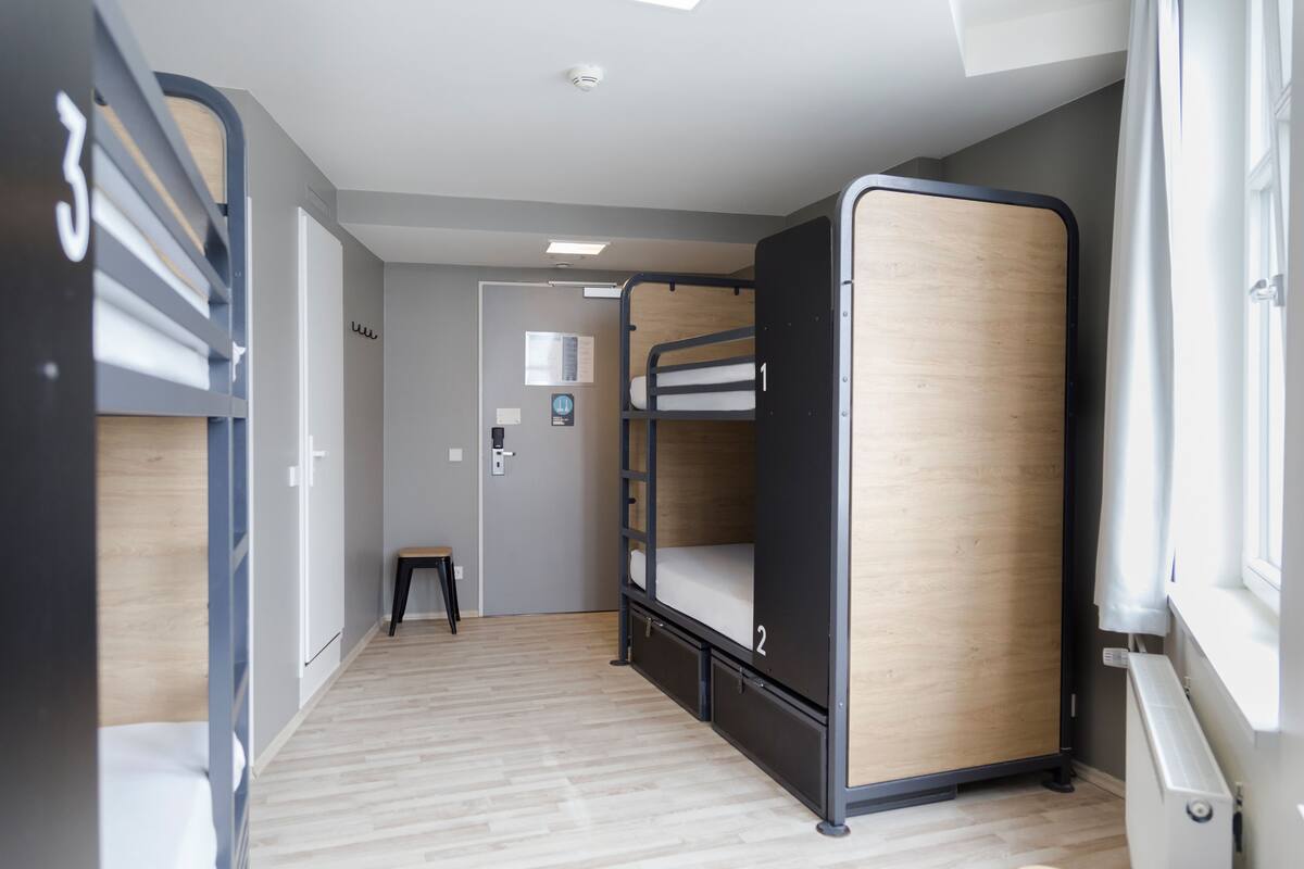 A room with bunk beds, a small stool, a radiator, and a door leading to another room.