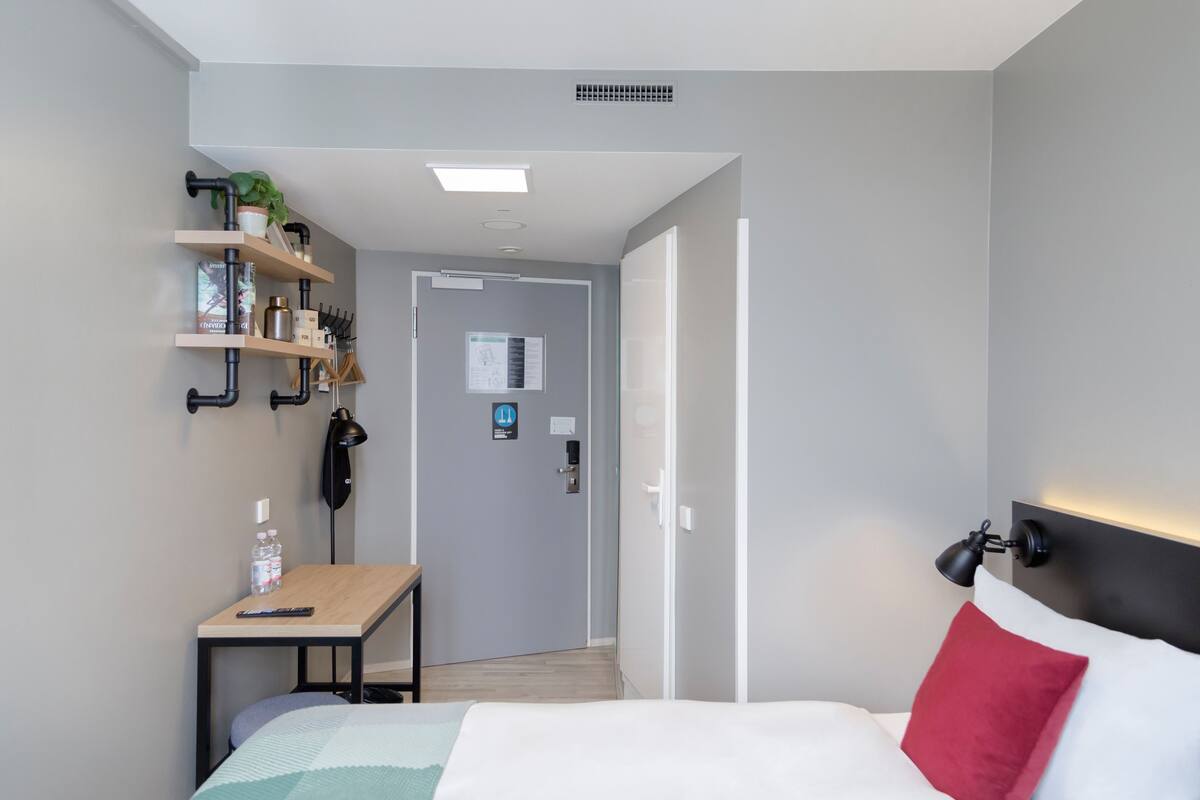 A hotel room with a bed, a desk, a shelf with toiletries, a door, and a wall-mounted lamp.