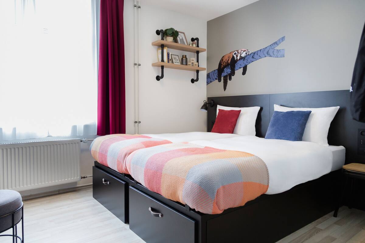 A modern bedroom with a large bed, a nightstand, a wall-mounted shelf with decorative items, and a cityscape view through the window.