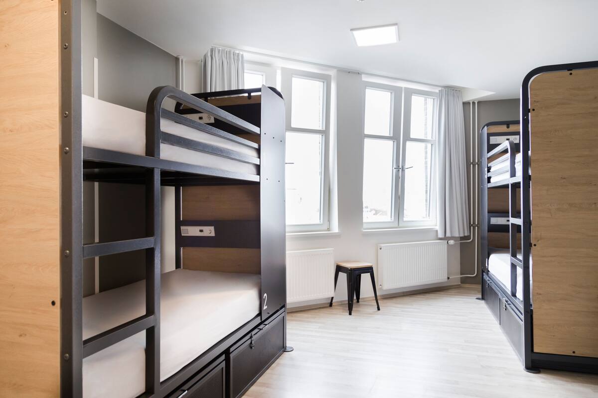 A dormitory room with bunk beds, a radiator, and a window with curtains.