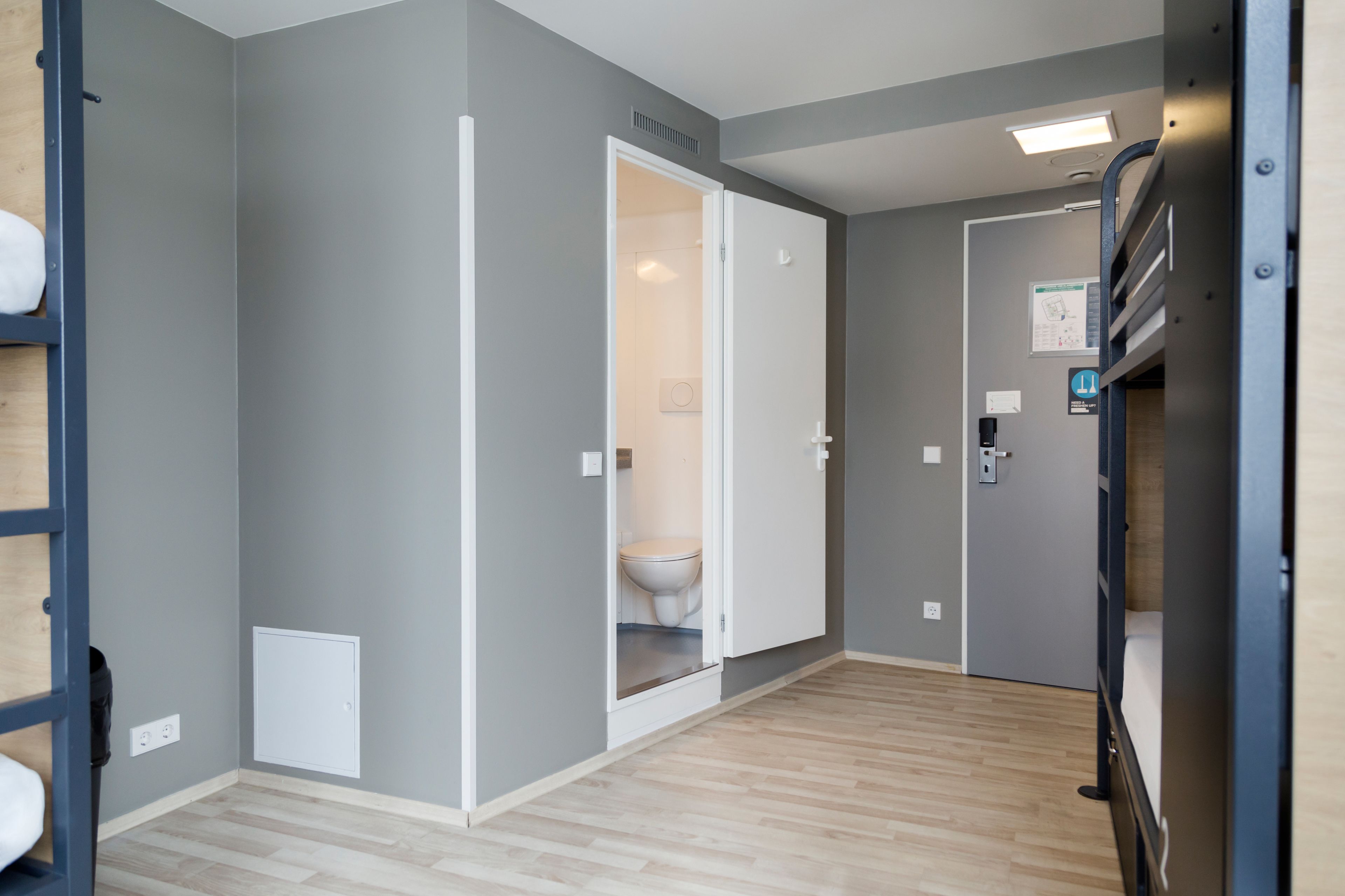 Un couloir avec un plancher en bois, une porte blanche donnant sur une salle de bain, et un lit superposé à l’arrière-plan.