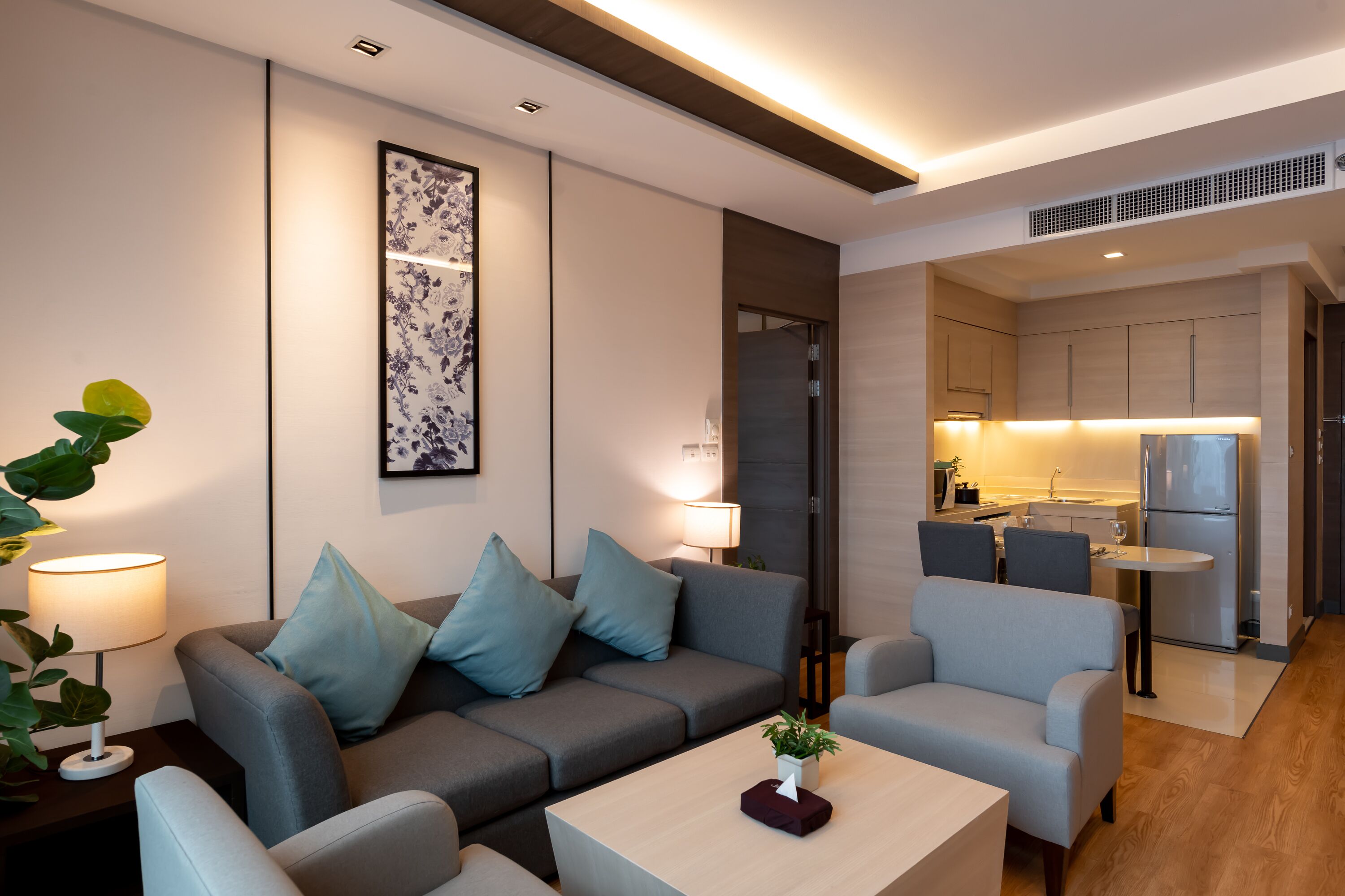 A modern living room with a sofa, armchairs, a coffee table, and a dining table. A kitchen is visible in the background with a refrigerator and cabinets.