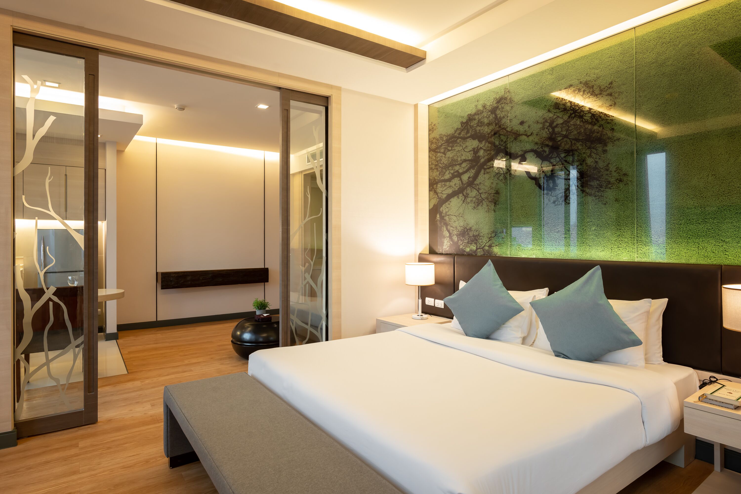 A modern hotel room with a large bed, a nightstand, a wall-mounted lamp, and a glass partition with a tree design.