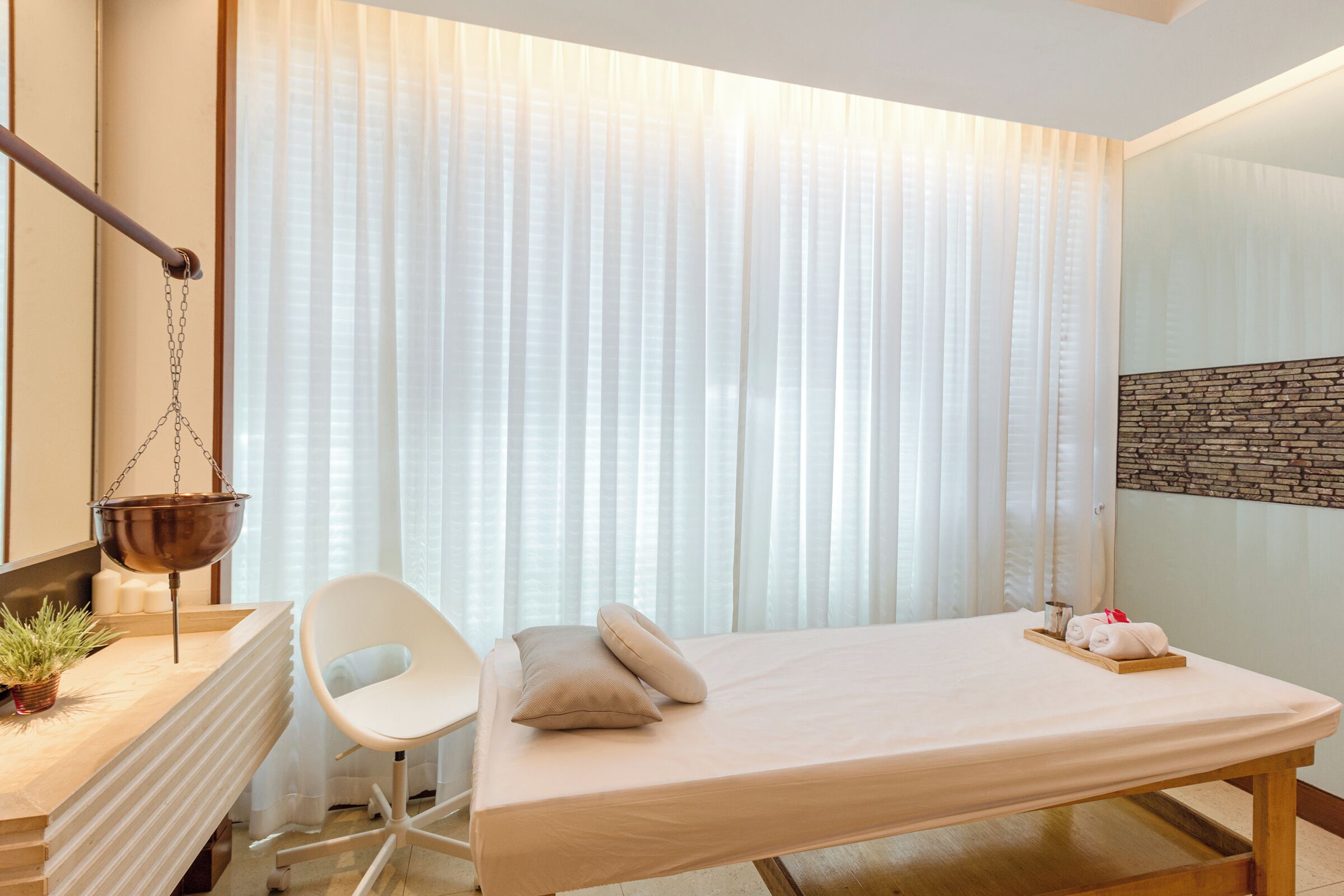 Un moderno spa con una camilla de masaje, una silla blanca, una bañera colgante y una pared de piedra.