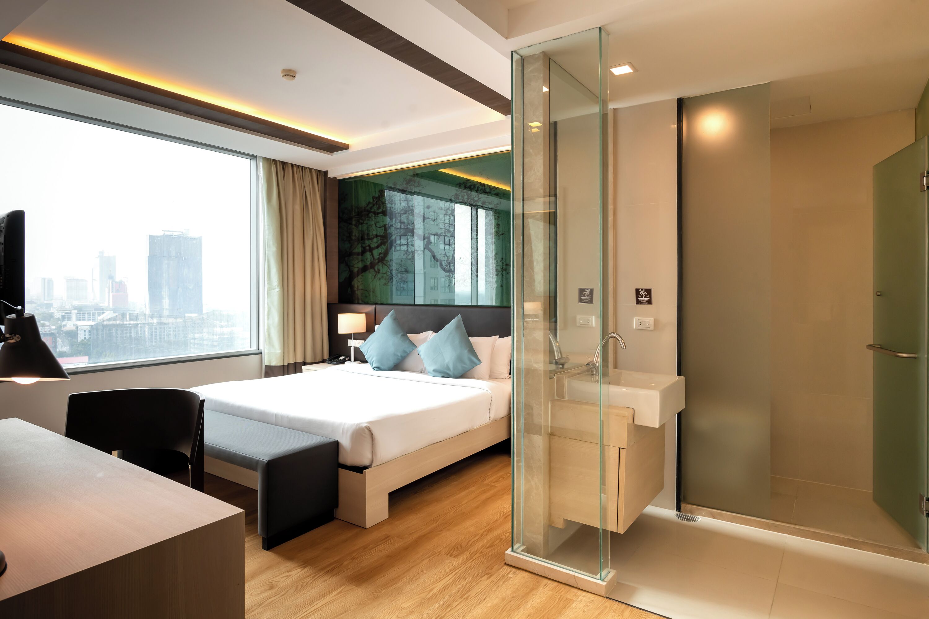 Una habitación de hotel moderna con cama, escritorio, silla, ducha envuelta en cristal y vistas de la ciudad.