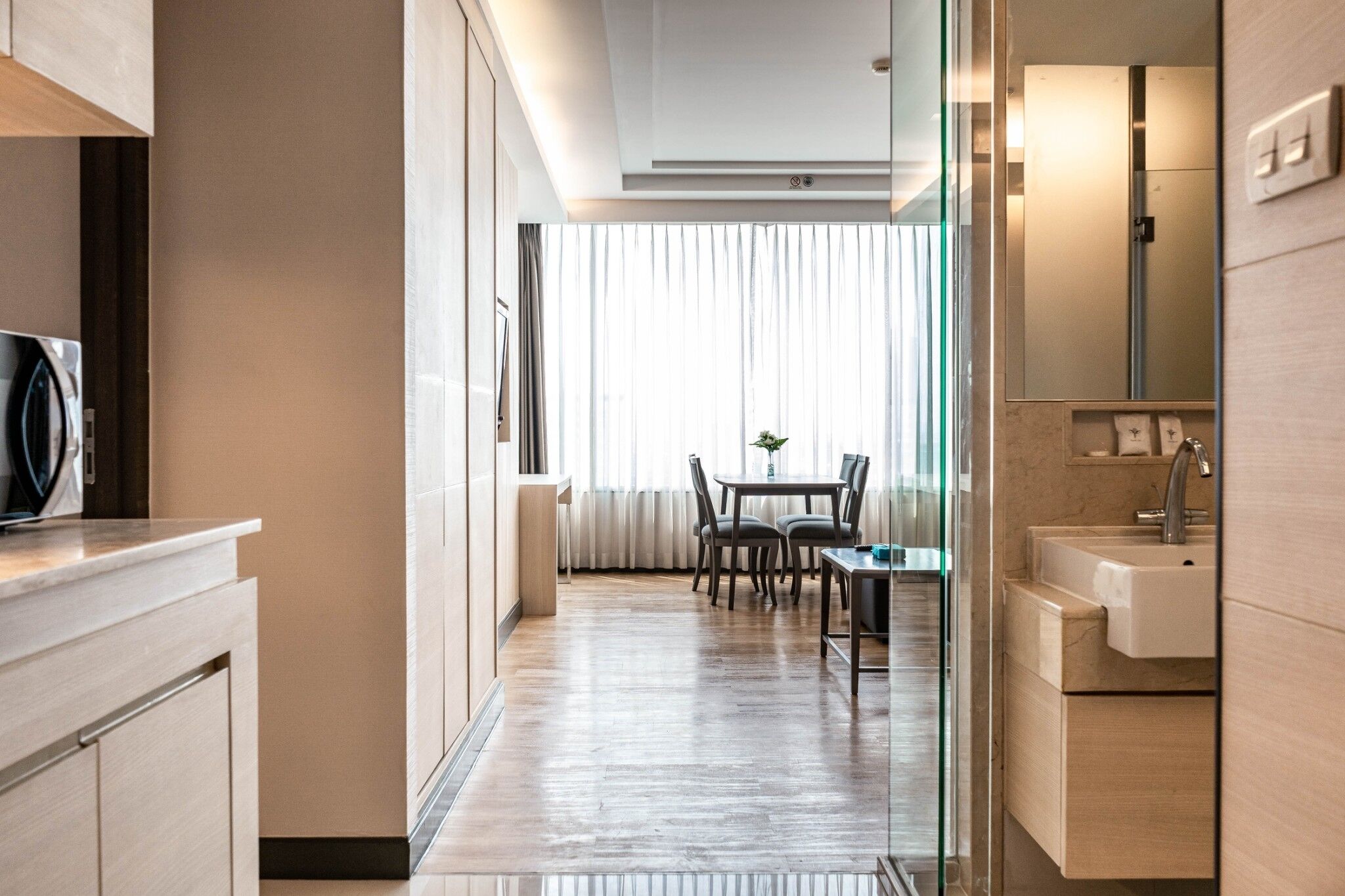 A modern hotel room with a wooden floor, a dining area with a table and chairs, a kitchen area with a sink and cabinets, and a large window with curtains.