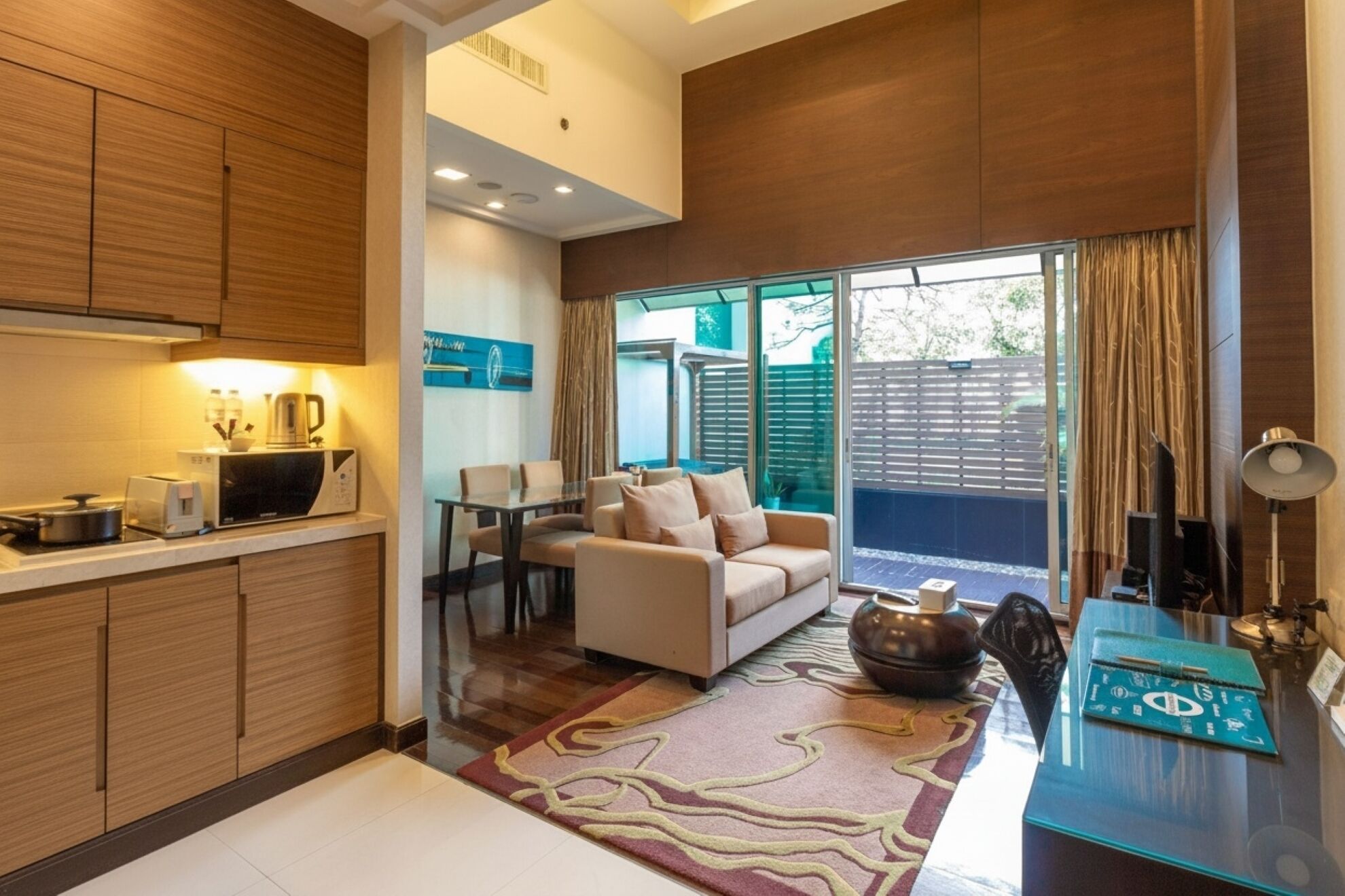 A modern kitchen with wooden cabinets, a dining area with a sofa and table, and a view of the outdoors through large windows.